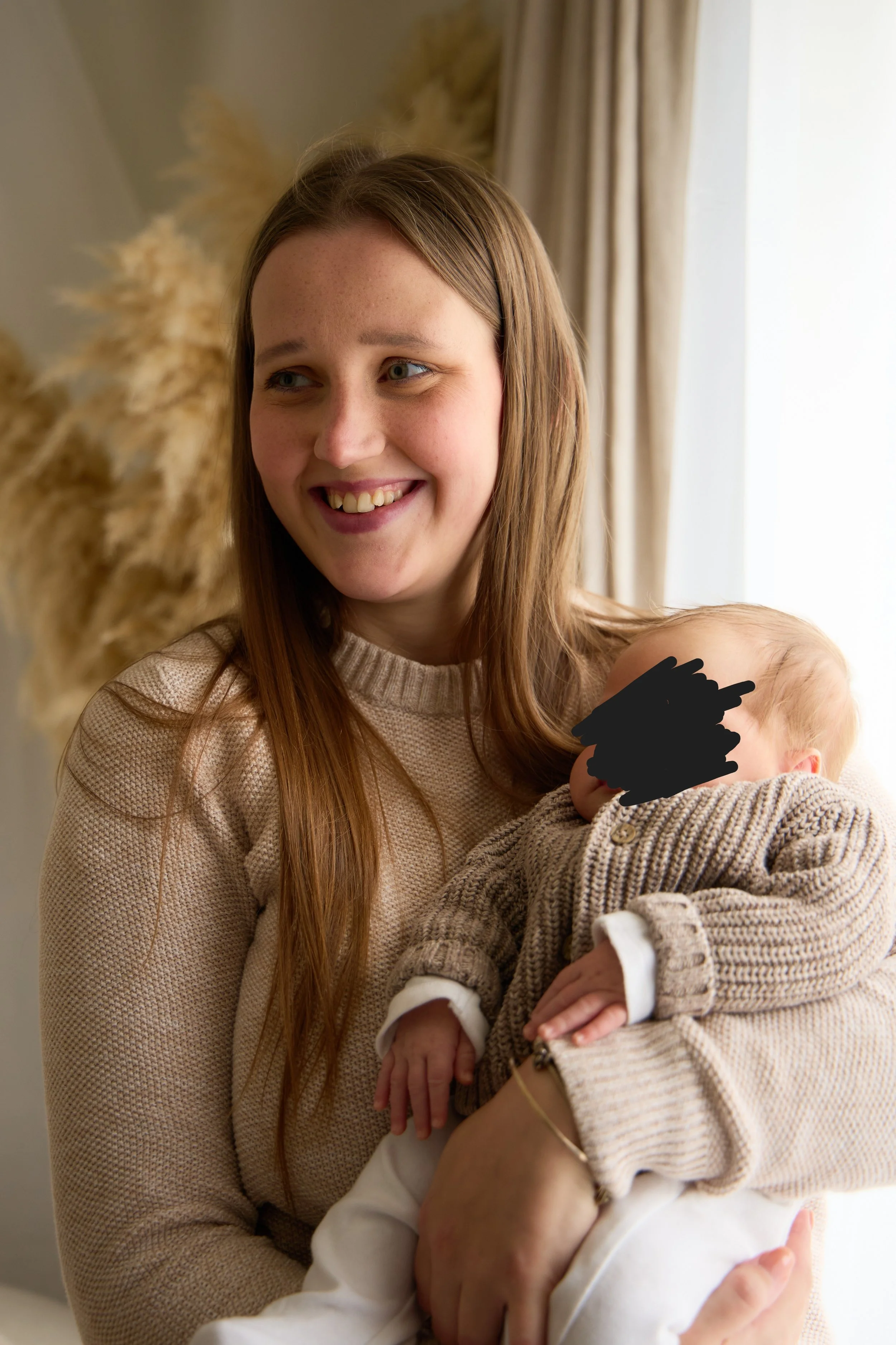 A woman holding a baby near a window with curtains and decorative dried plants in the background.