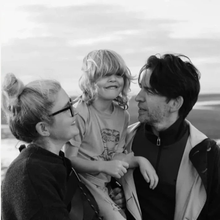 A woman, a man, and a young girl smile and look at each other on a beach. The woman and girl have blonde hair, and the man has dark hair. The woman wears glasses, and the scene is in black and white.