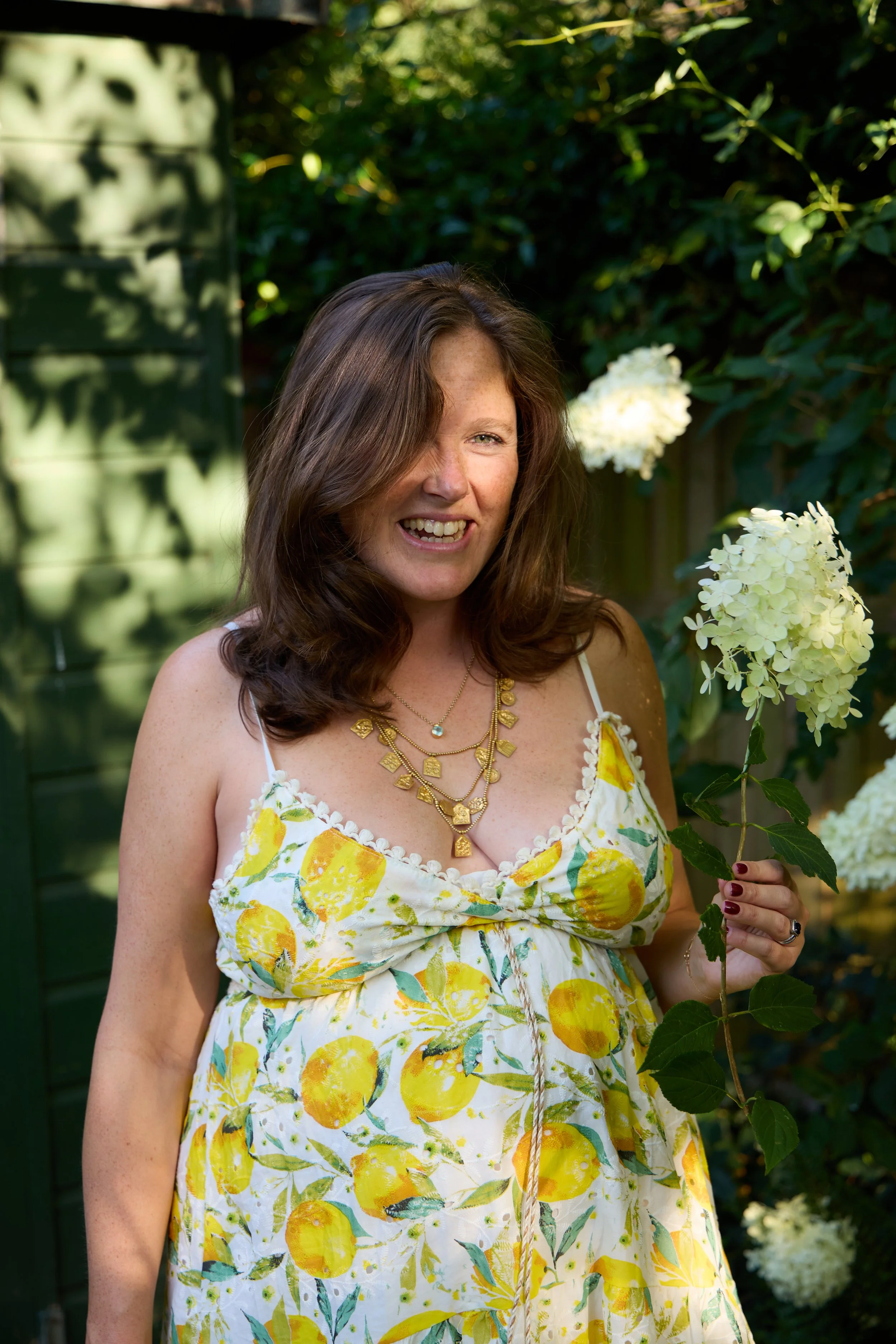 A woman with brown hair smiling and holding white hydrangea flowers while standing outdoors in a garden.