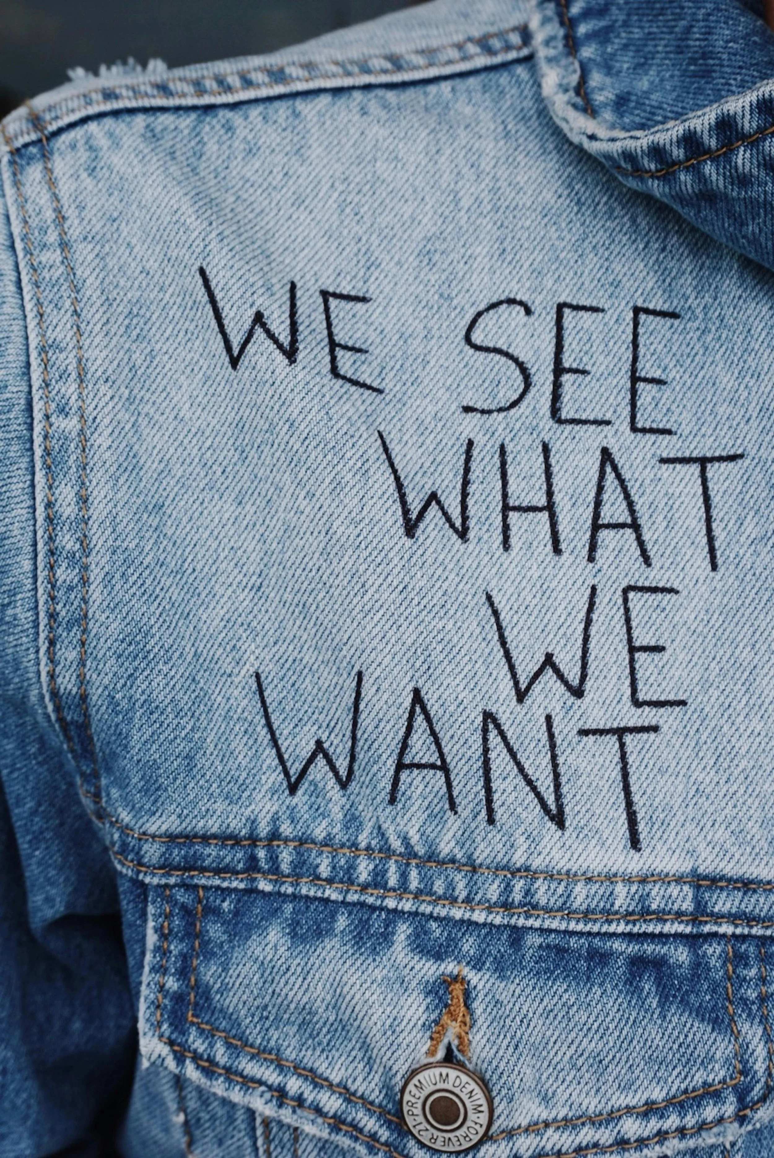 Close-up of a light blue denim jacket with black stitched text that reads 'WE SEE WHAT WE WANT'.