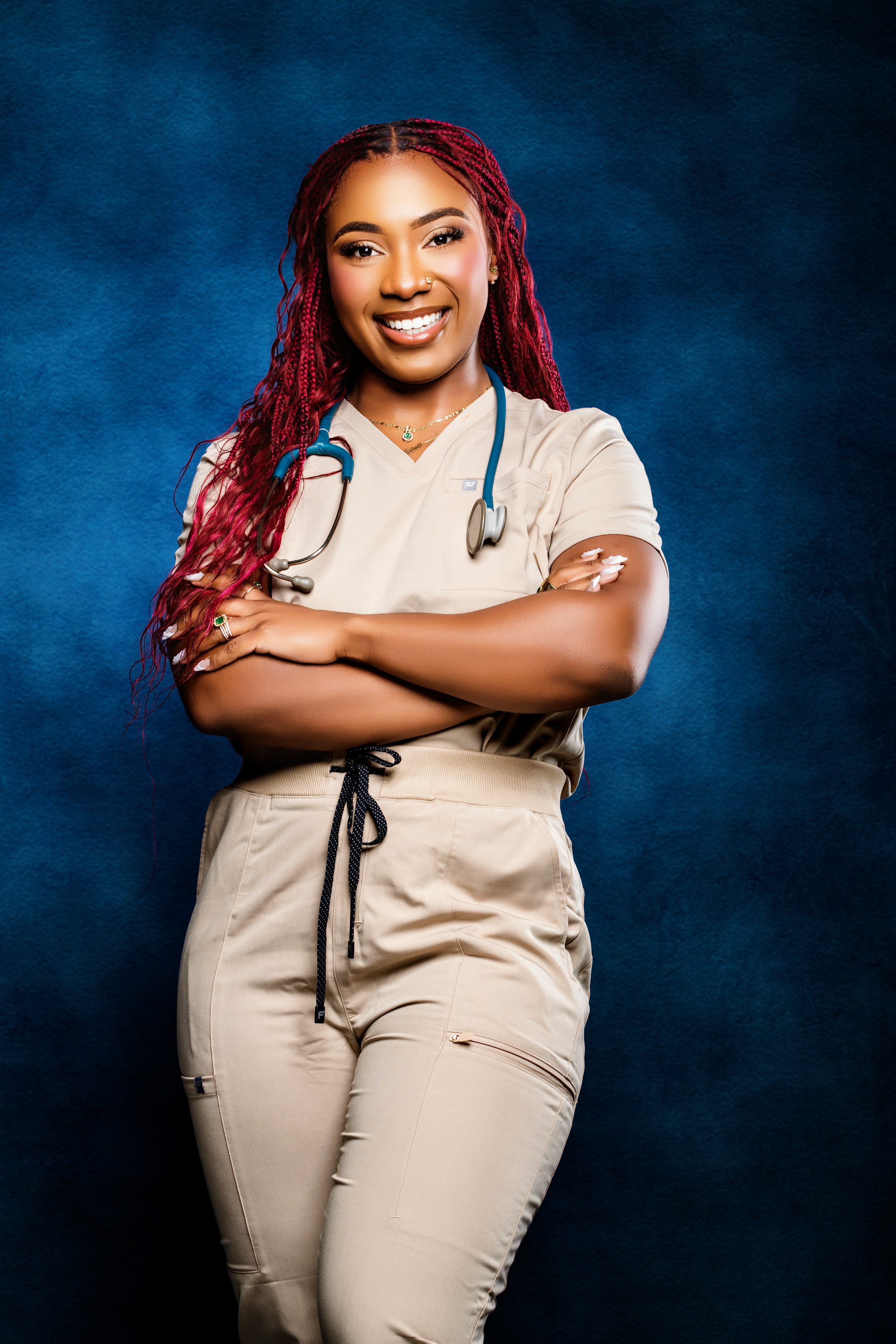 A smiling female healthcare professional wearing beige scrubs, with a stethoscope around her neck, standing with arms crossed against a dark blue background.