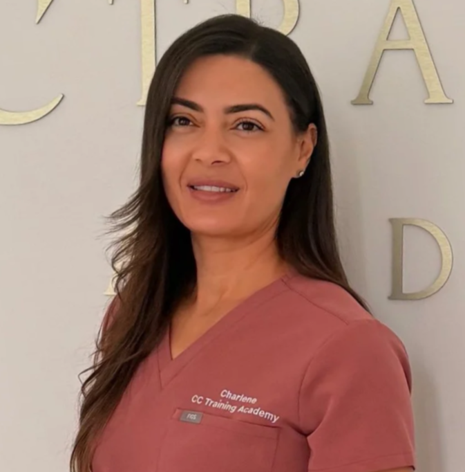 A woman with long brown hair wearing a pink uniform with the name tag 'Charlene' and 'CC Training Academy' standing in front of a wall with gold letters.