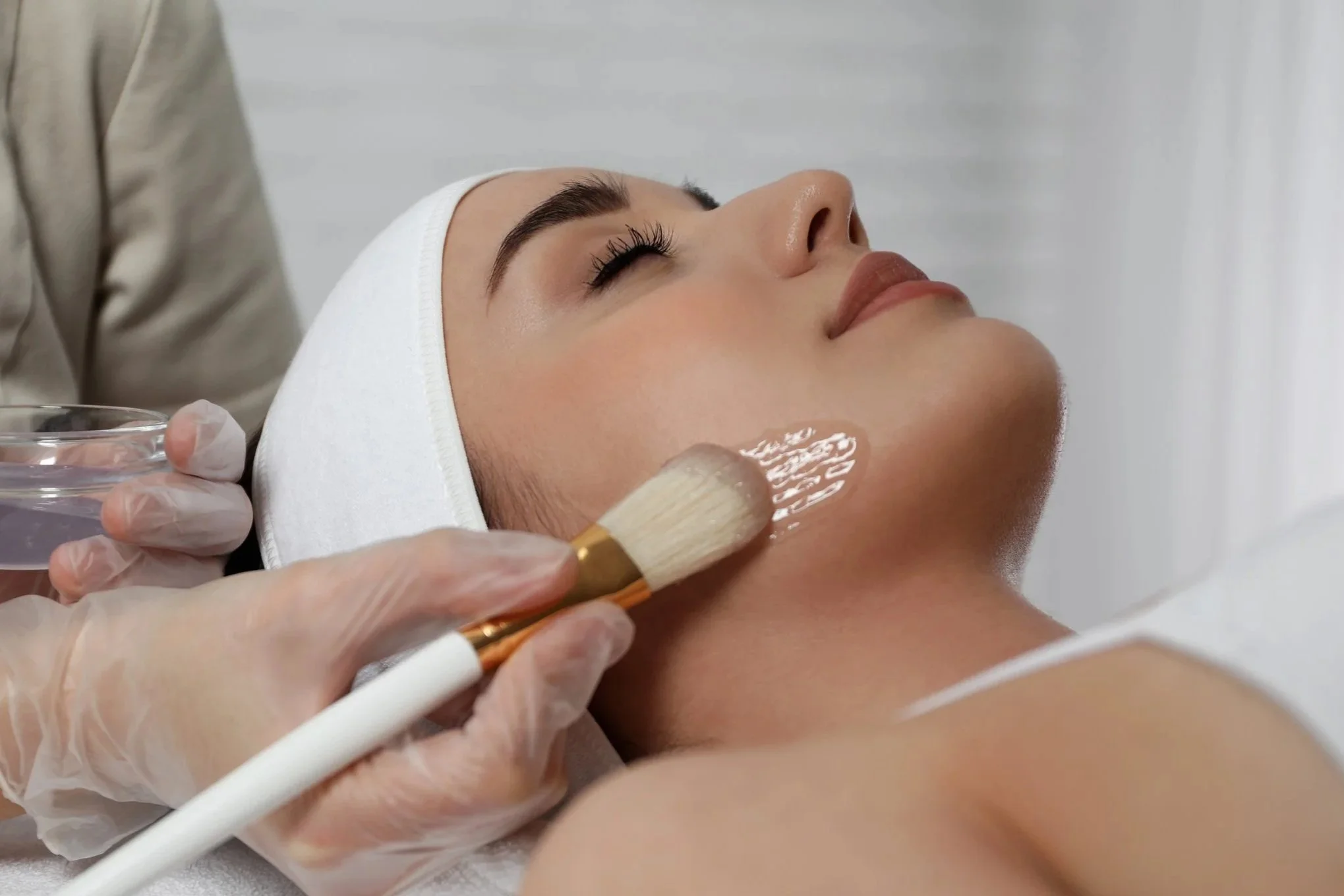 A woman receives a facial treatment from an esthetician, who is applying a skincare mask with a brush on her cheek while she has her eyes closed.