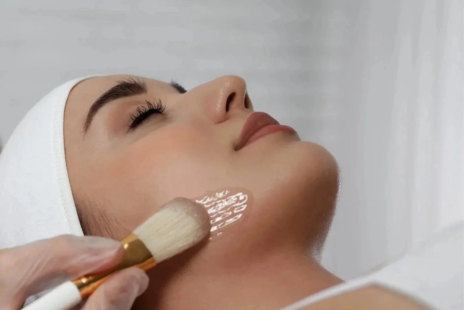 A woman receives a facial treatment. She is lying down with a white headband, and a skincare professional is applying a facial mask or cream with a brush.