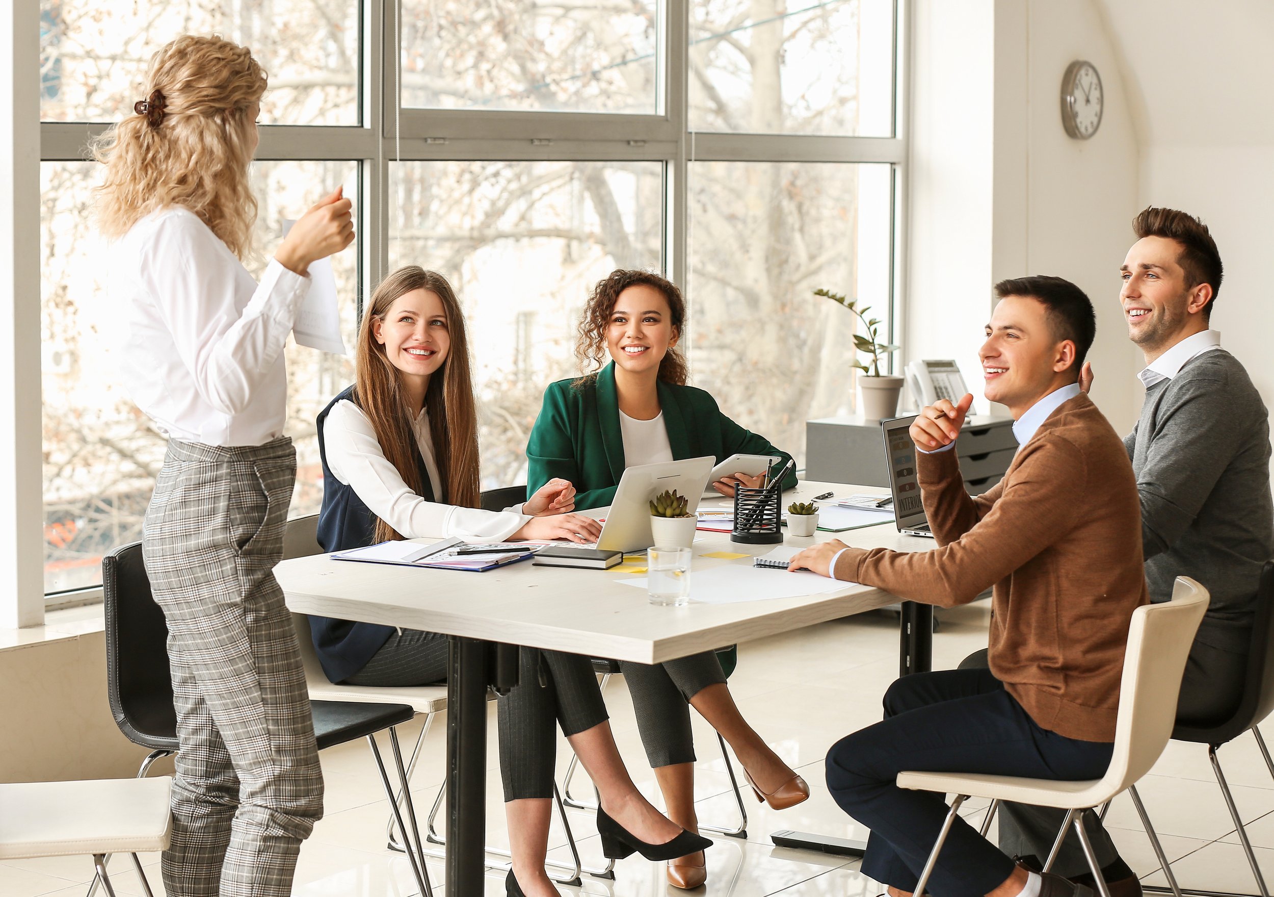 Five young professionals in a meeting room, engaged in a discussion. One woman is standing and speaking, while four others are seated at a table with laptops and notebooks, smiling and listening.