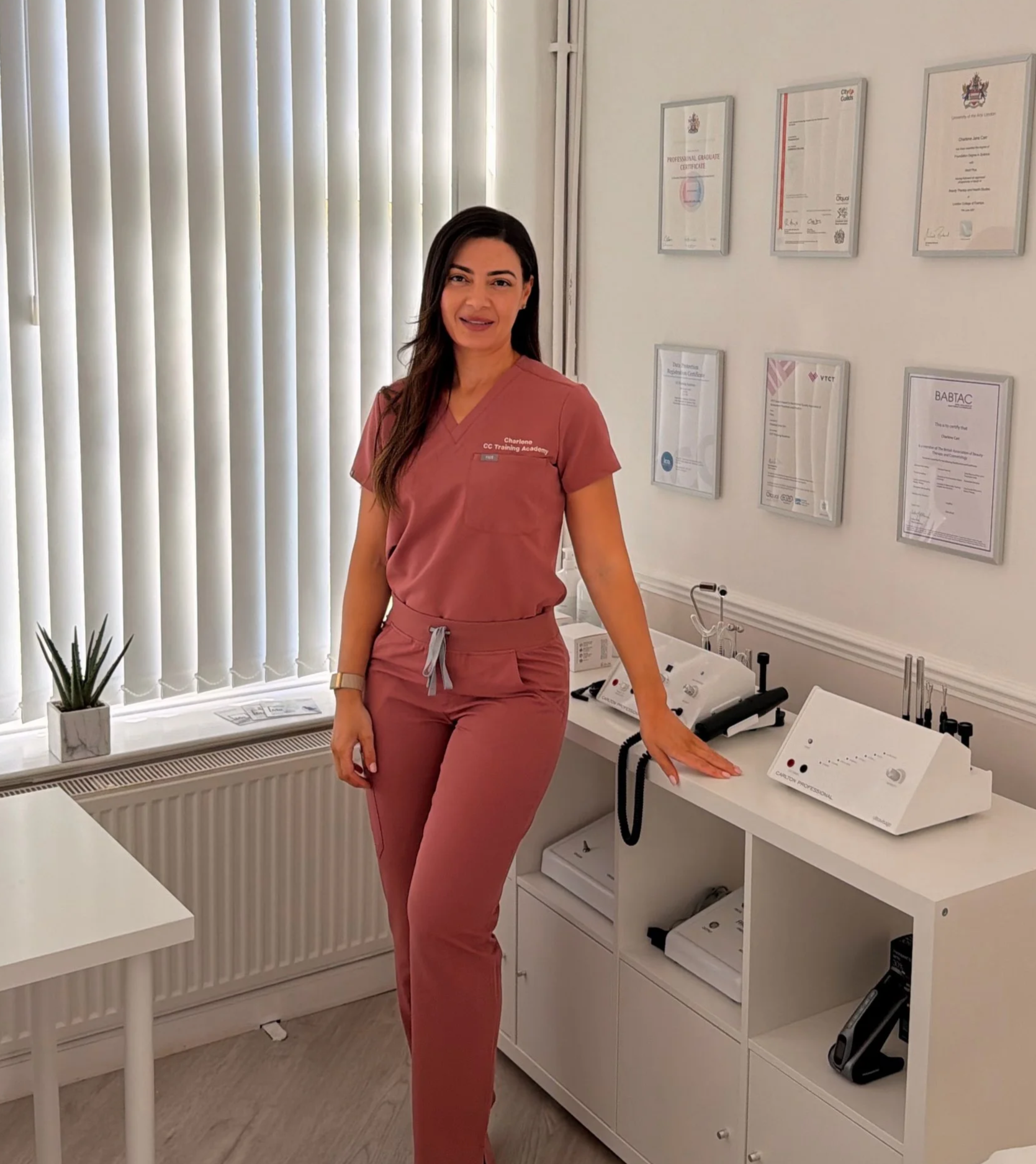 A woman wearing pink medical scrubs stands in a clinic or office setting, with a white cabinet and framed certificates or diplomas on the wall behind her. She is smiling slightly and leaning on a counter with medical equipment nearby.