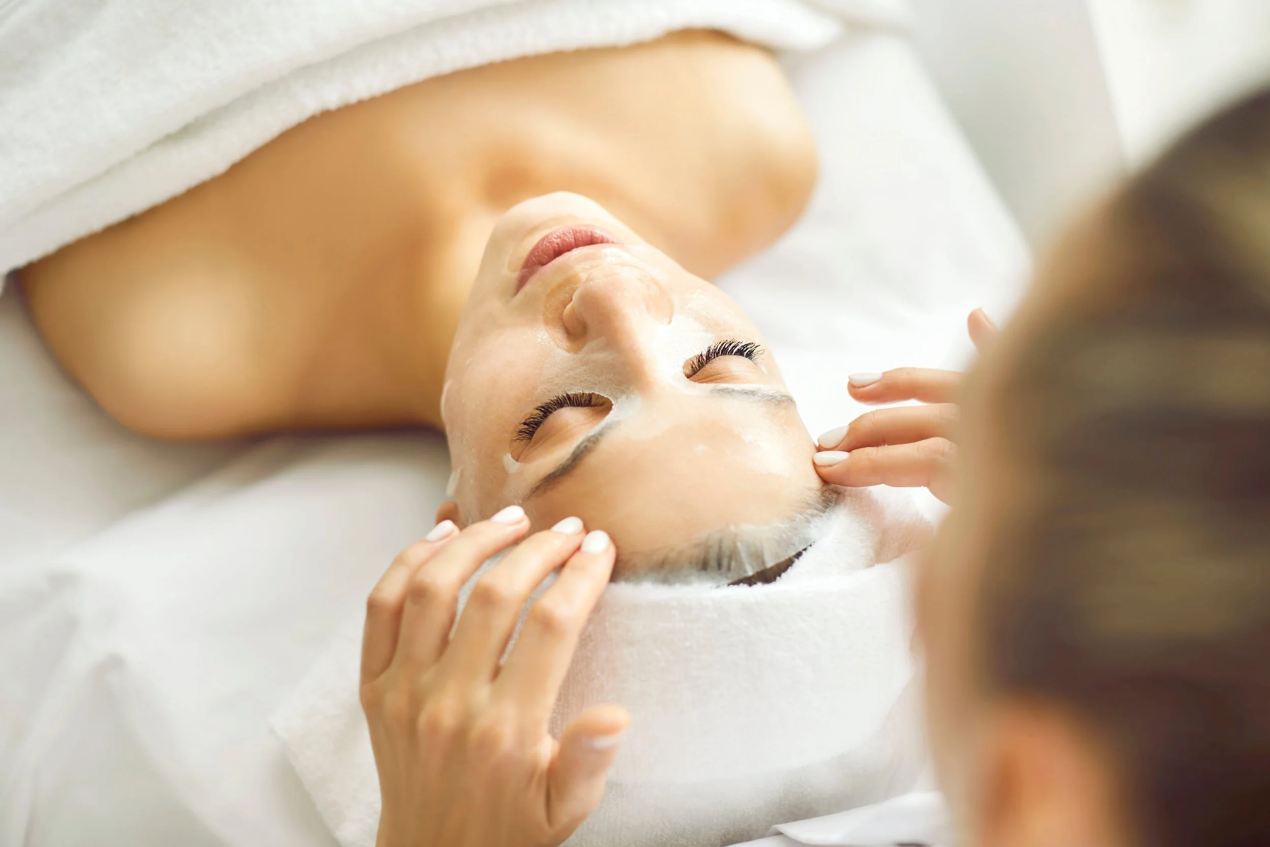 A woman receiving a facial treatment at a spa, lying on a bed with her eyes closed, with a beautician gently touching her forehead.