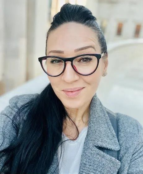 A woman with dark hair, wearing glasses and a gray coat. She is smiling and taking a selfie indoors.