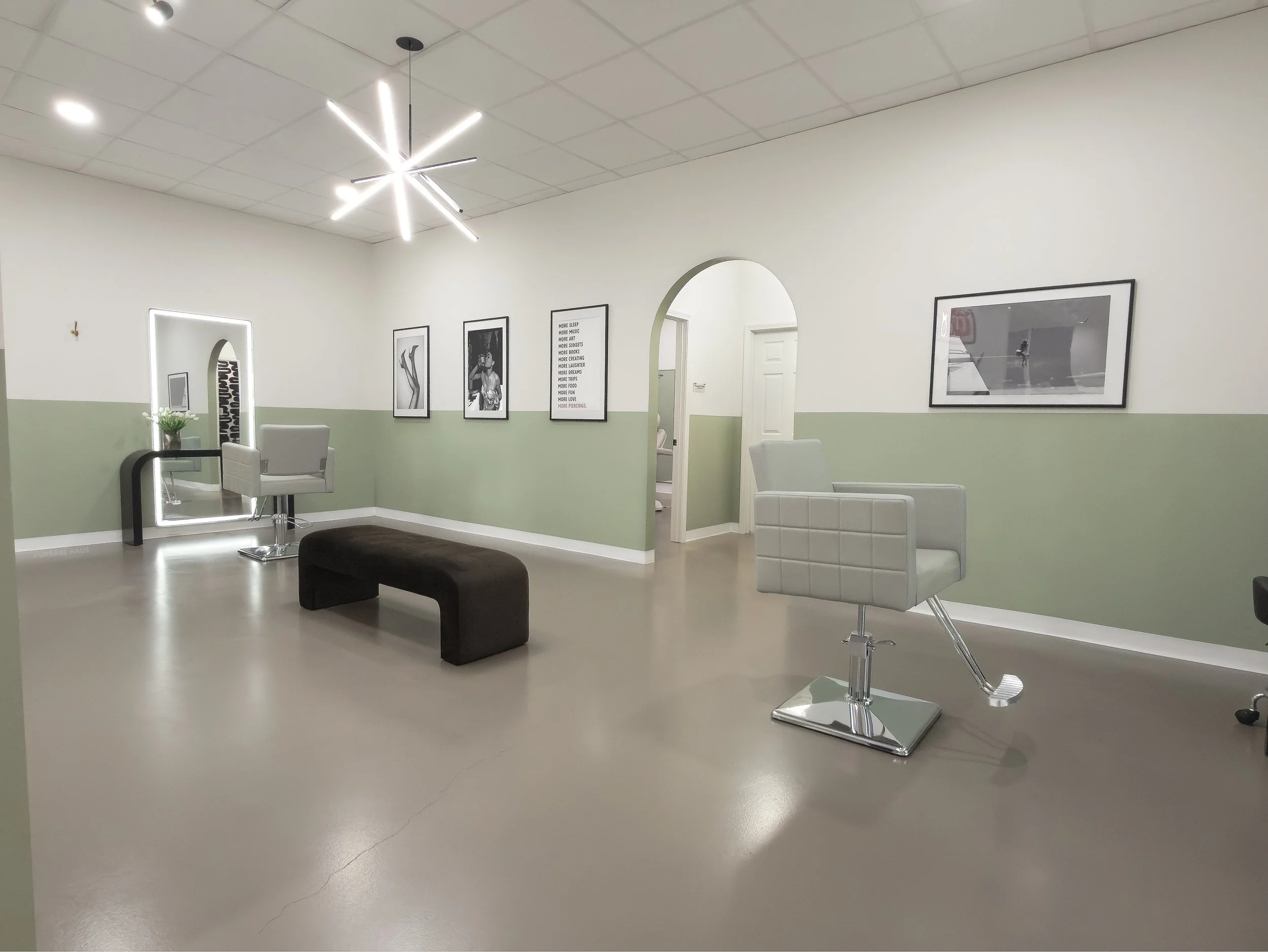 Empty hair salon with styling chairs, a black bench, wall art, chandelier, and a mirror with flowers on it.