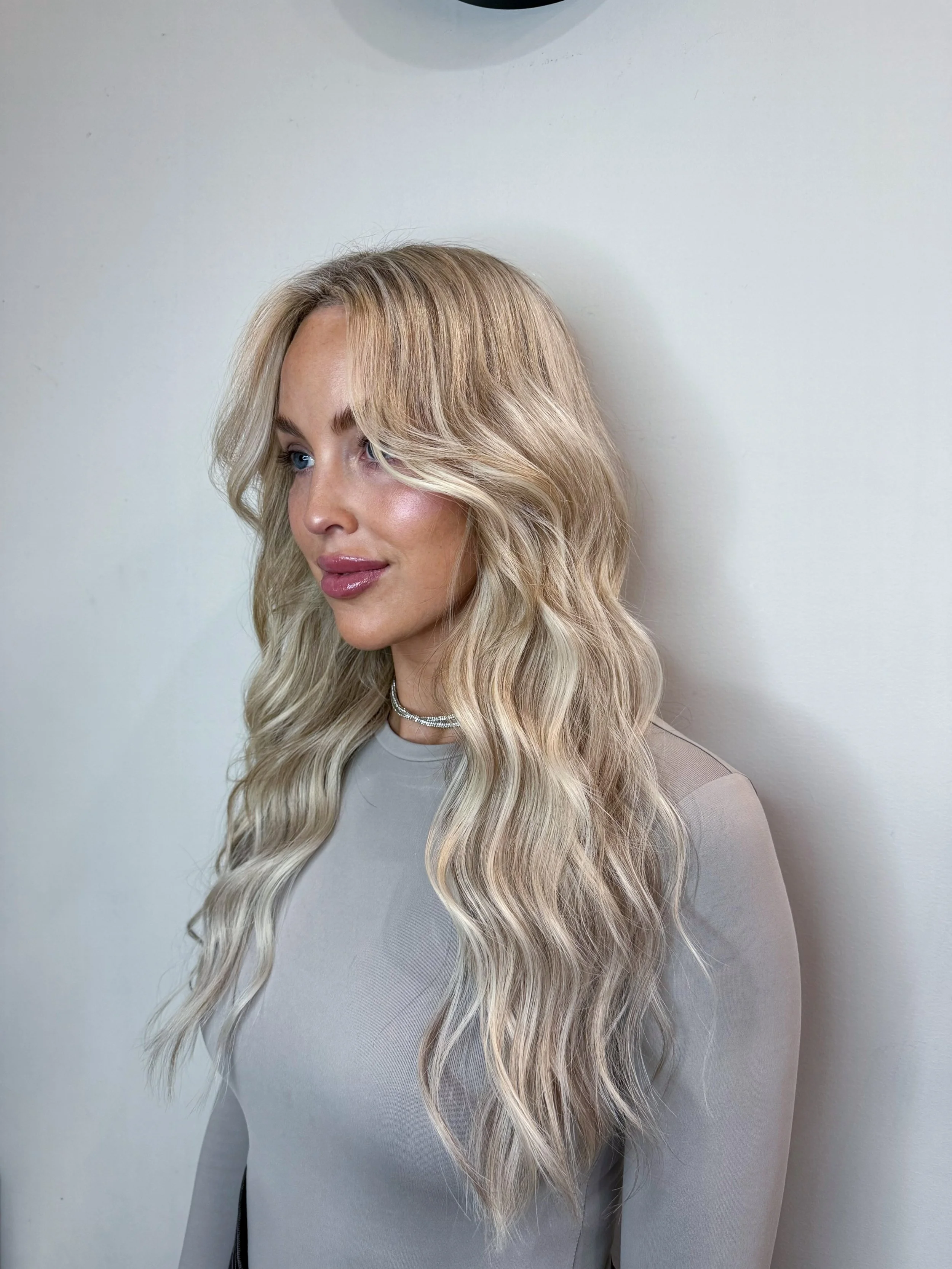A woman with long, wavy blonde hair, wearing a light gray top and a silver necklace, standing against a plain white wall.
