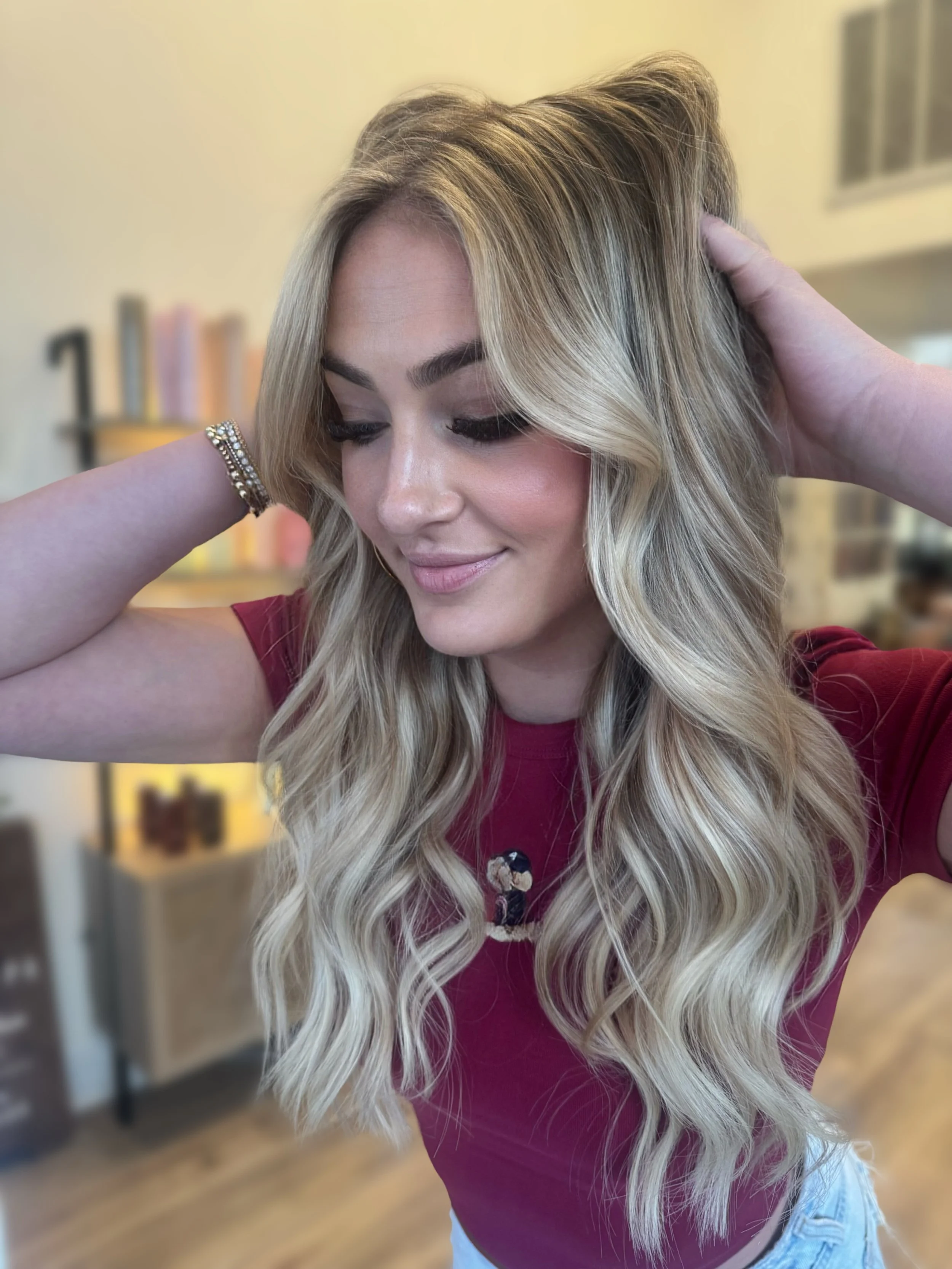 A woman with long, wavy blonde hair, wearing a maroon shirt and a bracelet, smiling with her eyes closed, in a salon or home setting.