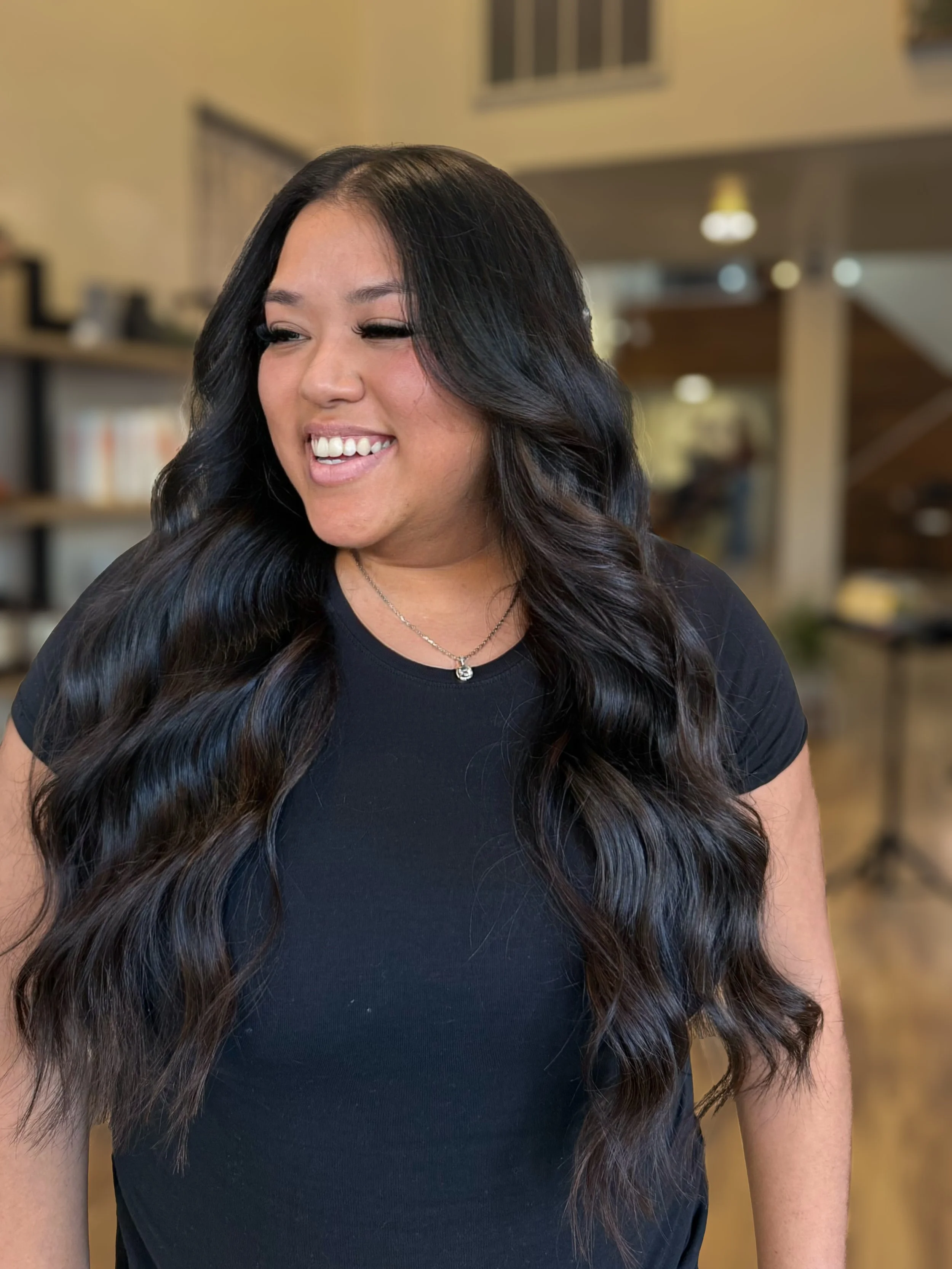 A woman with long, wavy black hair smiling and turning her head to the side in a cozy indoor space.