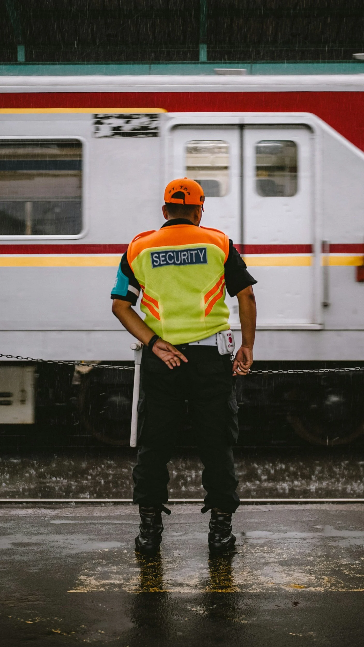 Security guard standing on wet pavement in front of a train during rain, facing away from the camera, wearing a fluorescent yellow jacket with 'SECURITY' patch.