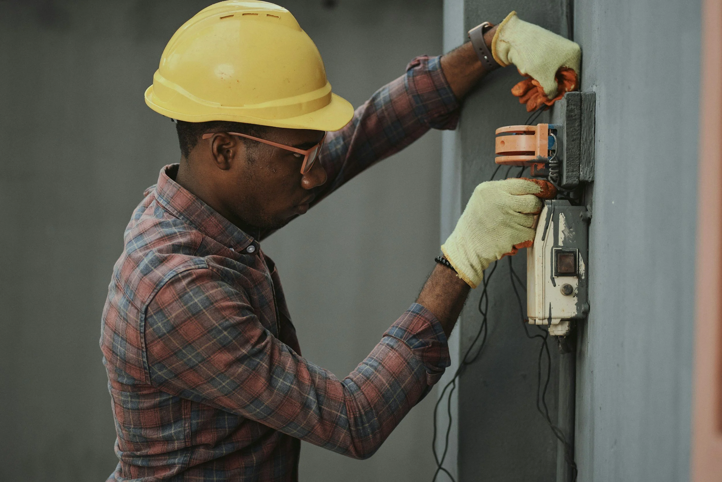 A man wearing a yellow hard hat, glasses, and yellow gloves working on electrical wiring on a gray wall, using a screwdriver.