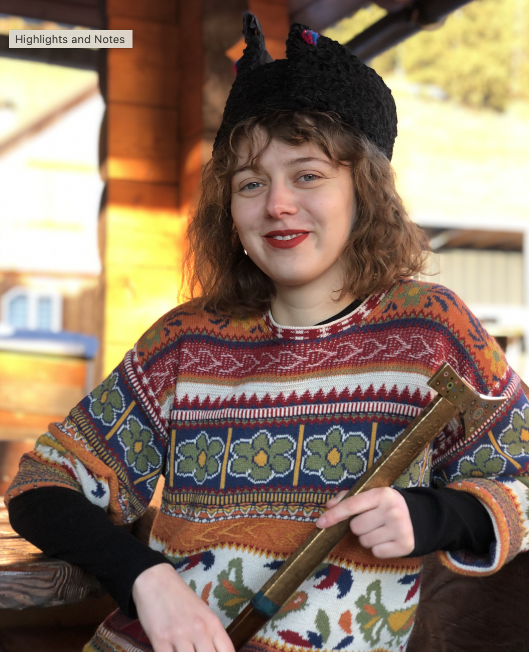 A young woman with curly red hair wearing a colorful, patterned sweater and a black furry hat with pink and blue accents. She has red lipstick and is holding a gold-colored hammer, smiling at the camera. She is outdoors in a setting with warm lighting and a wooden structure in the background.