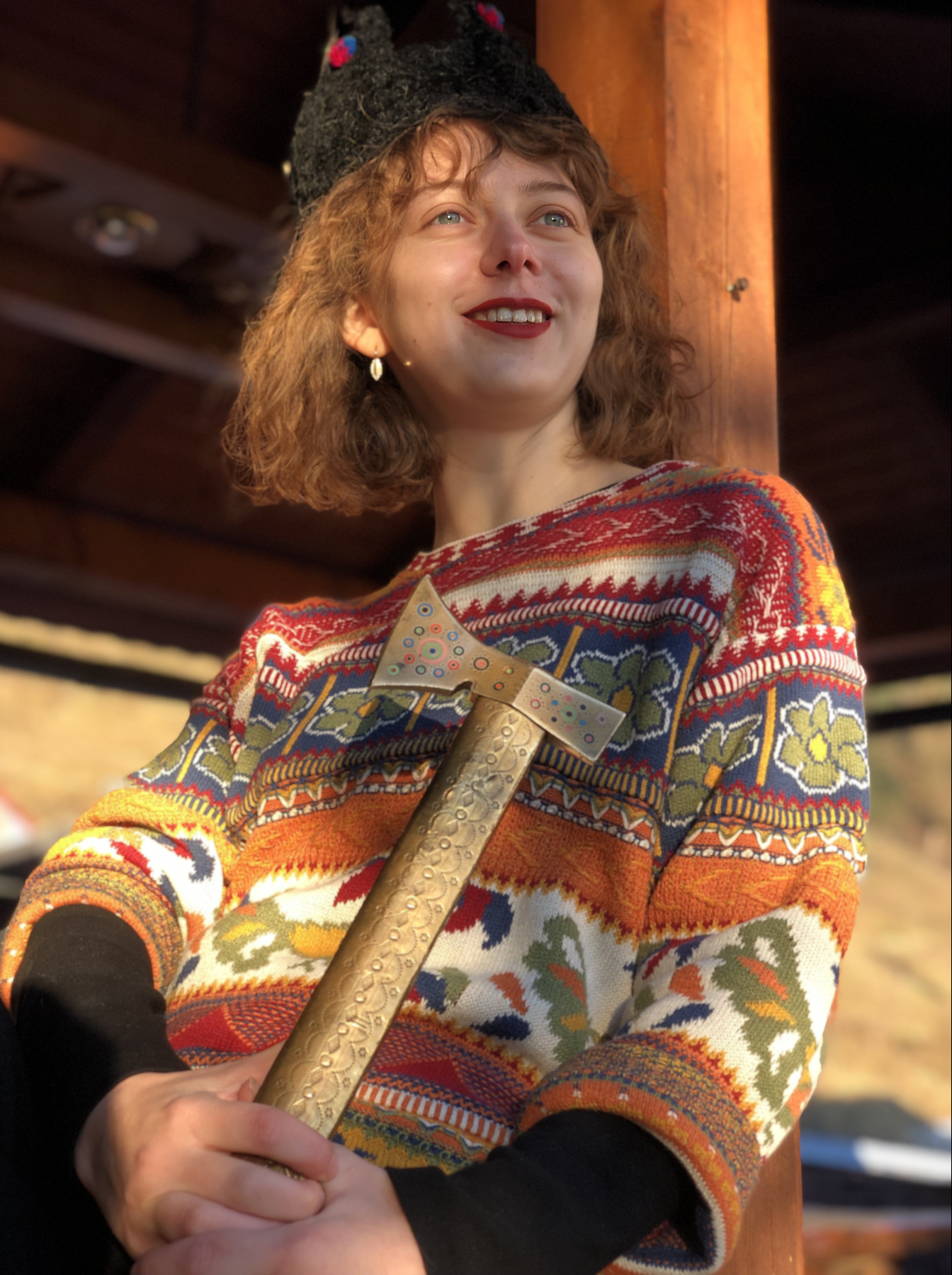 A woman with curly red hair and blue eyes, smiling and looking slightly to her right, holding a golden hammer. She is wearing a colorful, patterned sweater with shades of red, orange, yellow, blue, green, and white. The background appears to be a wooden structure, possibly part of a cabin or outdoor setting.