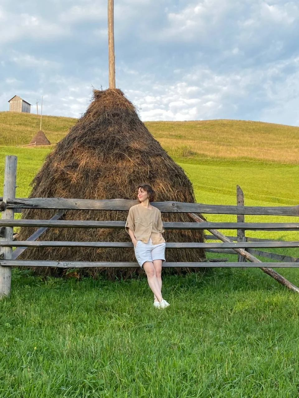 A woman in beige jacket and white shorts leaning against a wooden fence, with a large haystack behind her in a green field under a partly cloudy sky.