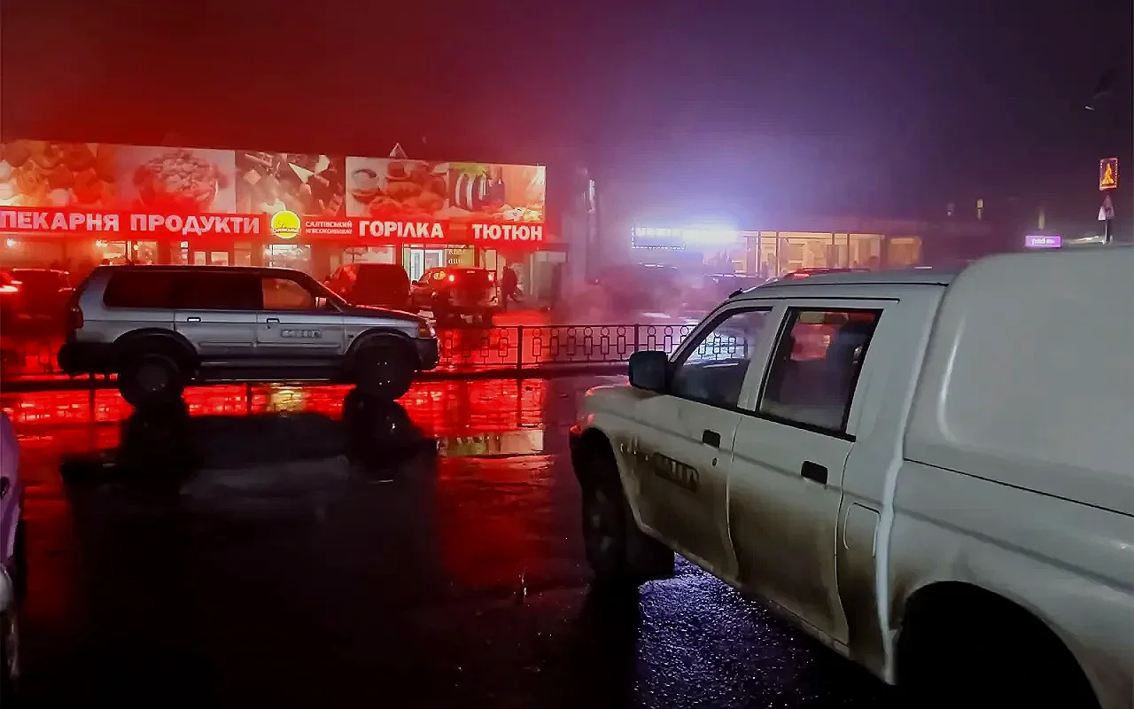 Nighttime scene of a wet parking lot with several cars, including a white van, in front of a storefront with illuminated signs in a non-English language. Red and blue lights create reflections on the wet surface.