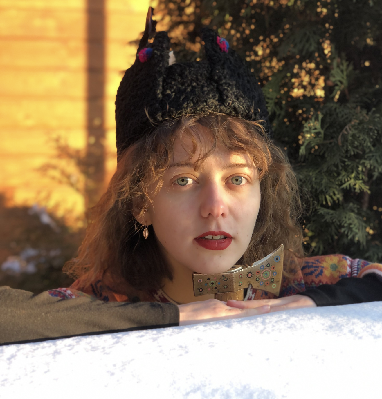 Young woman with wavy brown hair, blue eyes, wearing red lipstick, earrings, a colorful sweater, black gloves with a long sleeve, a black hat with pink accents, and a bow tie necklace, leaning on a white surface outdoors with trees and warm light in the background.