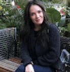 A woman sitting outdoors on a wicker chair with greenery in the background.