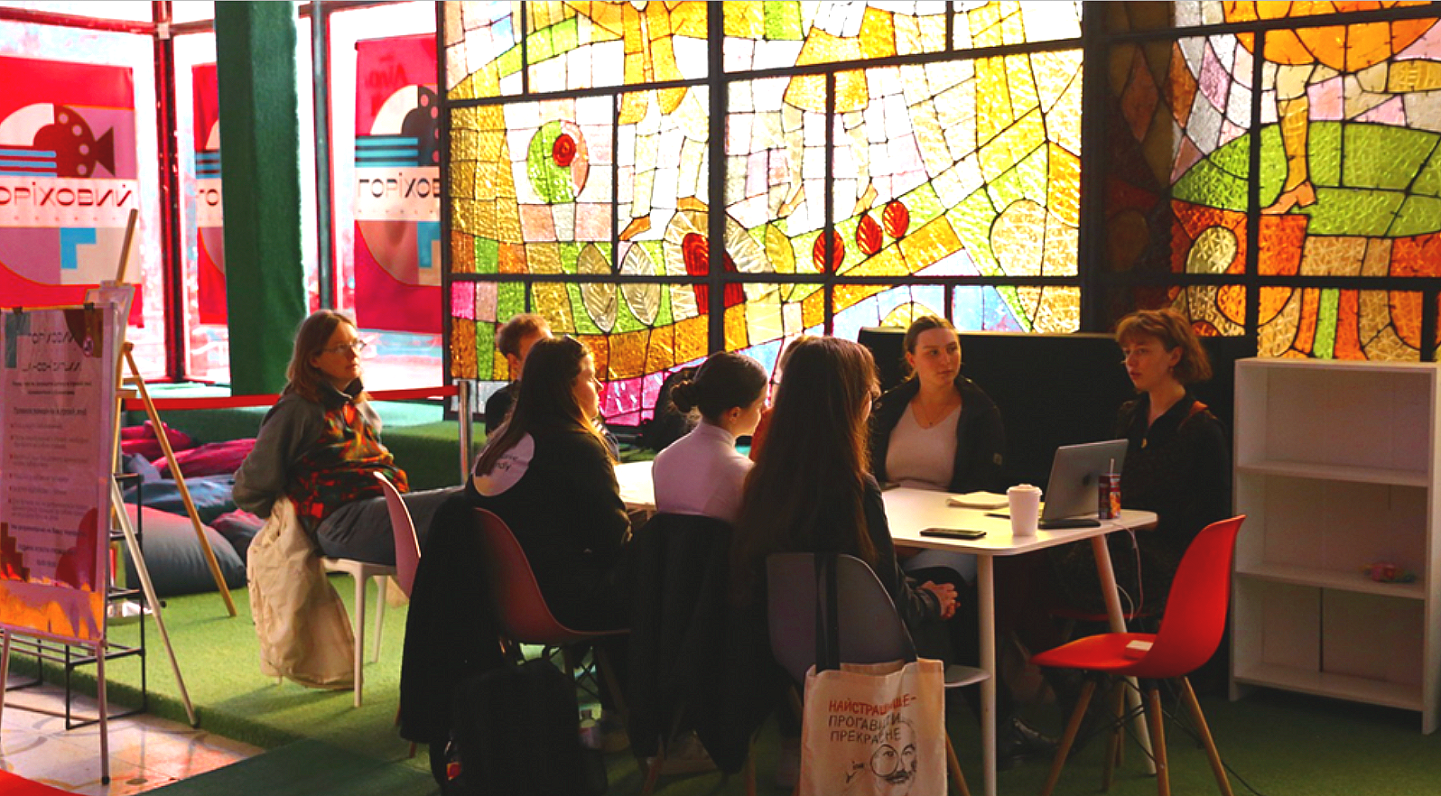 A group of people sitting around a table in front of a large mosaic stained glass window in a colorful, indoor space.