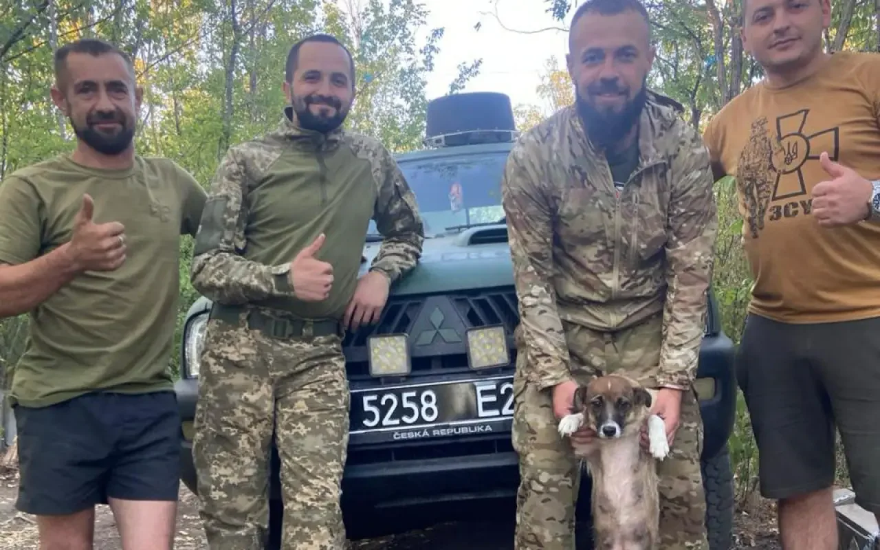 Four men sunglasses standing in front of a green military vehicle in a forest, one holding a small brown and white dog.