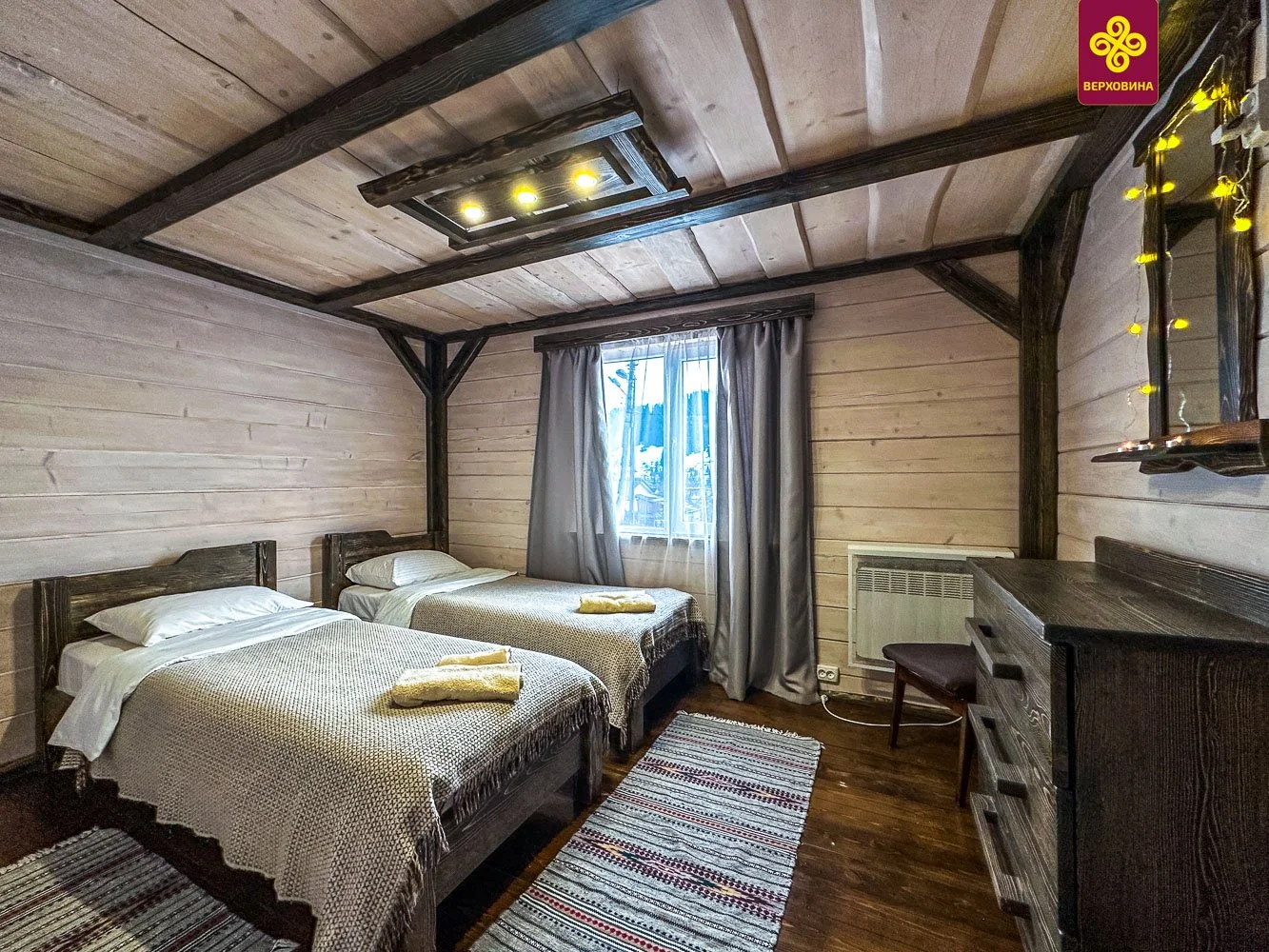 A rustic bedroom with two single beds, dark wood furniture, striped rugs, a window with curtains, and wooden paneled walls and ceiling.