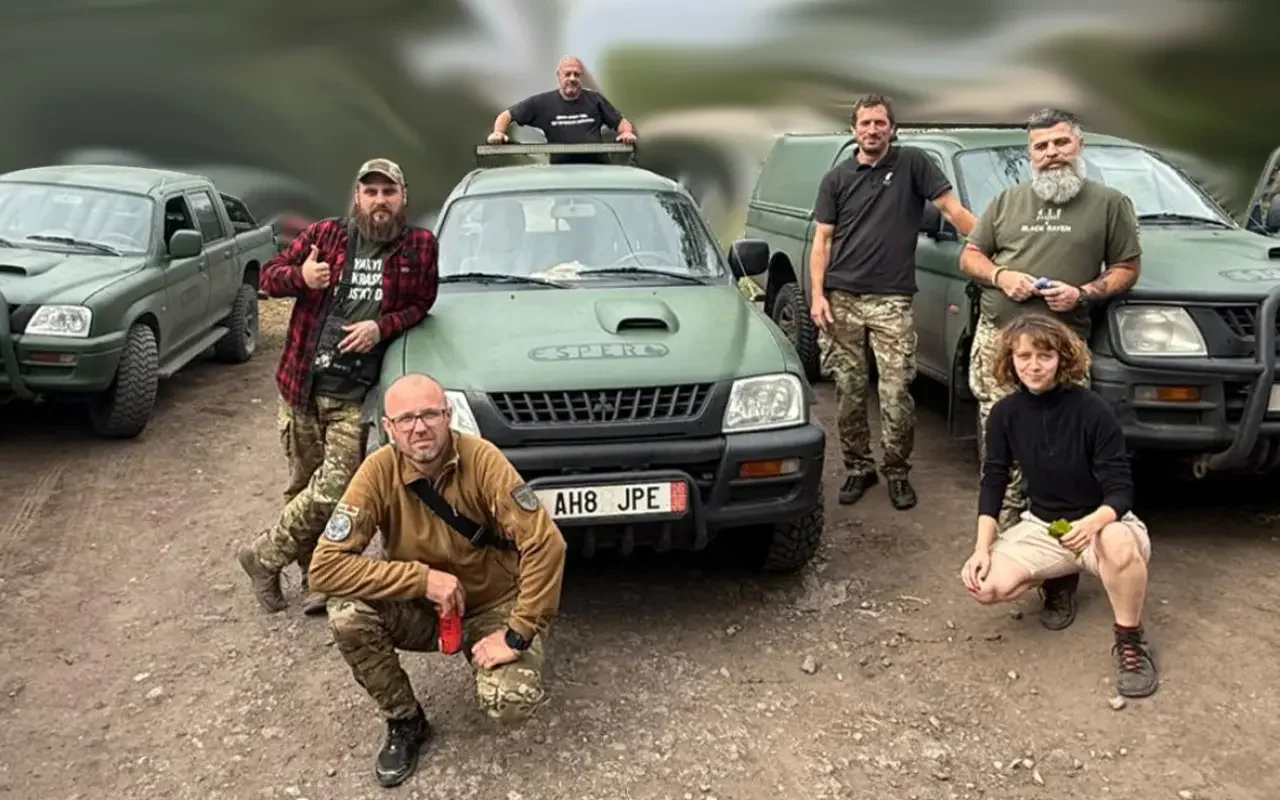 Group of six people, four men and two women, standing and kneeling in front of military-style vehicles on a dirt ground.