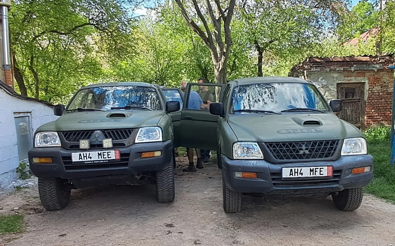 Two dark green Mitsubishi off-road vehicles parked side by side on a dirt path under tree canopy, with four people standing near the open doors, in front of a rustic brick and stone building.