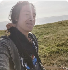 A woman standing outdoors on a grassy hill with a cloudy sky in the background.