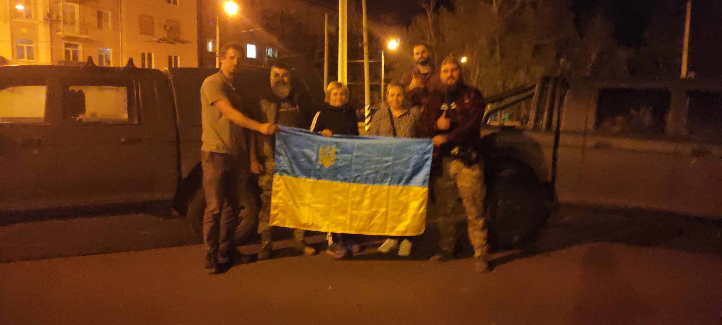 Group of six people holding a Ukrainian flag in front of a dark vehicle at night, with streetlights and buildings in the background.