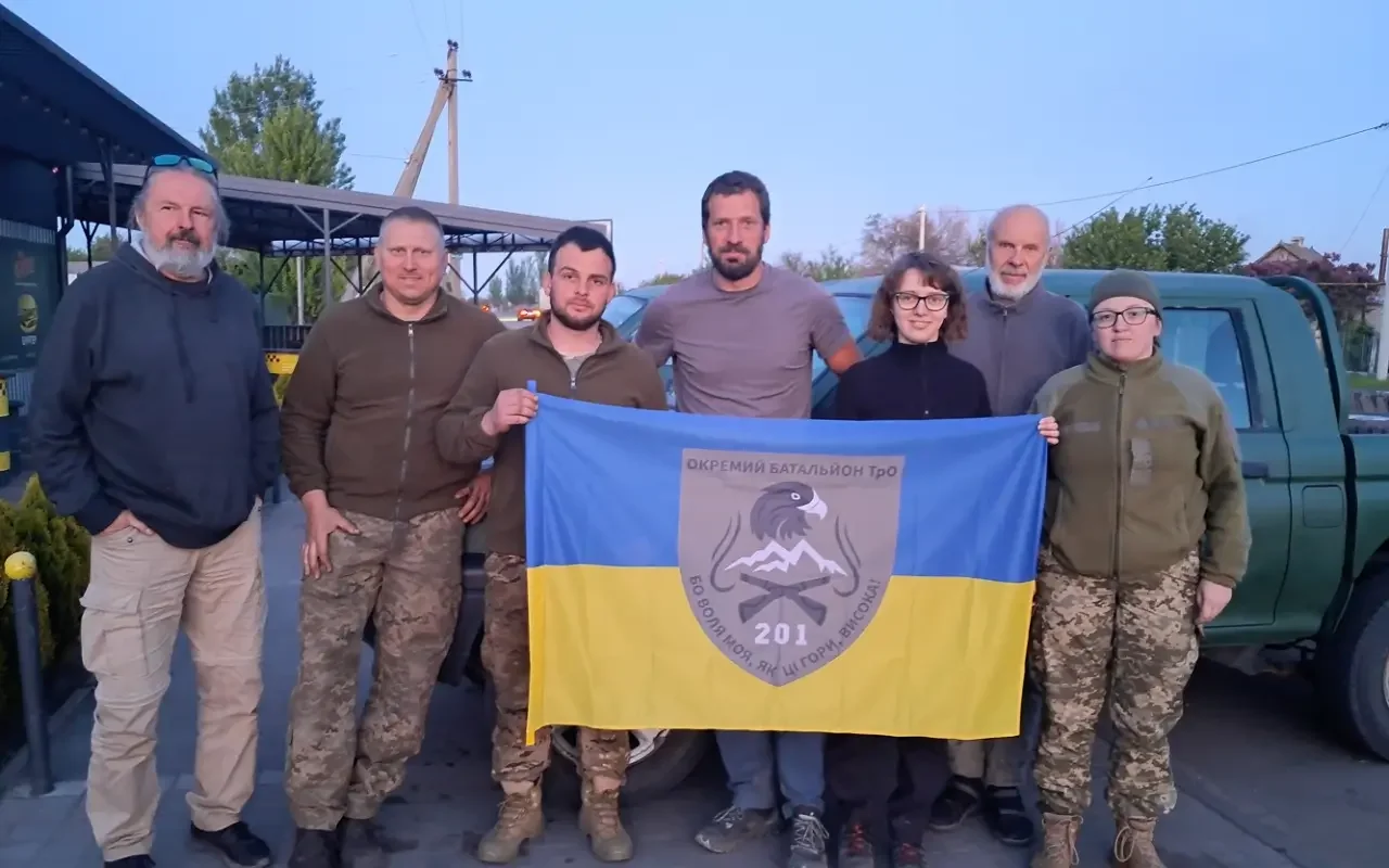 Group of seven people holding a blue and yellow flag with a crest, standing outdoors near cars and a building.