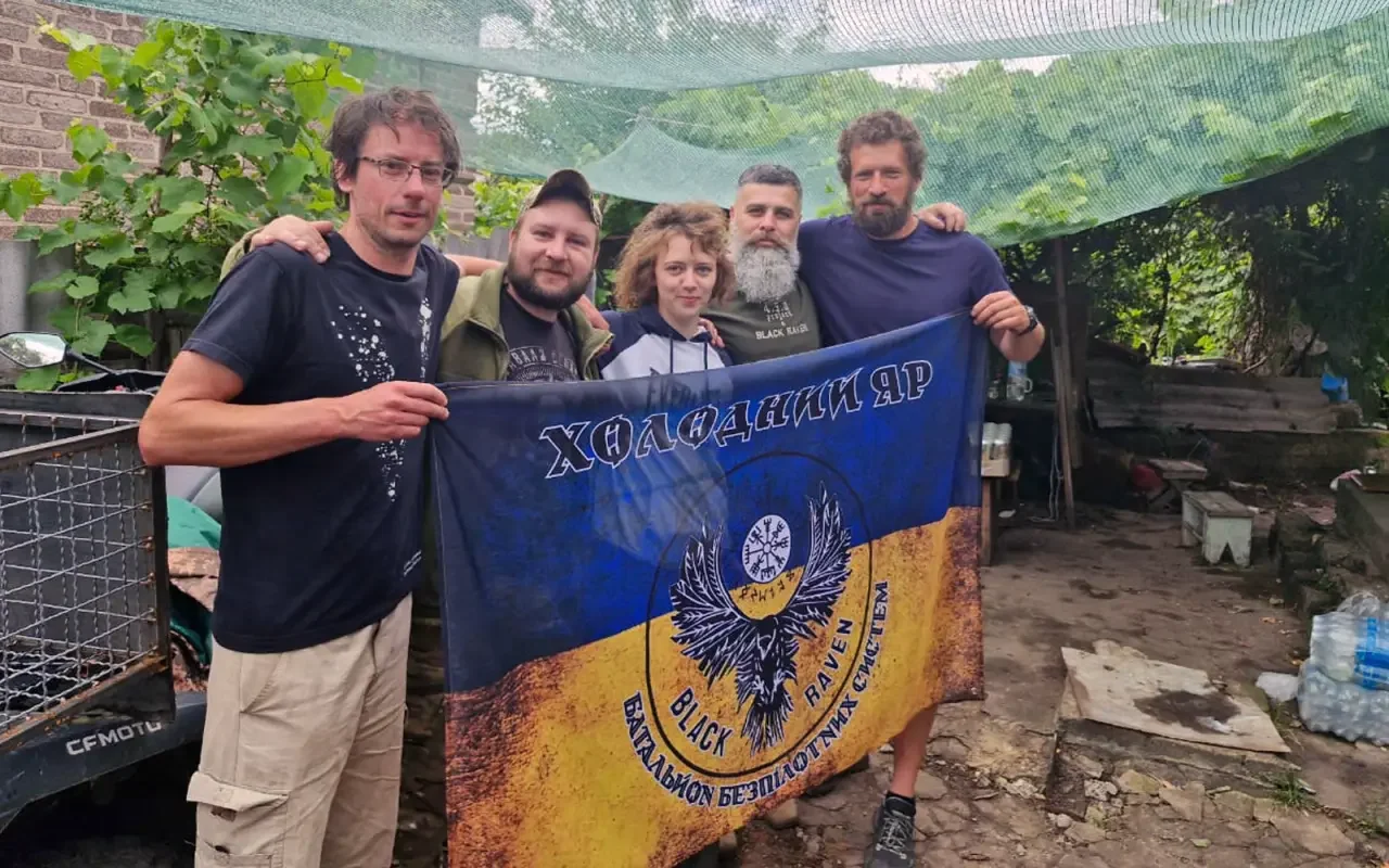 Five people standing outdoors under a green canopy holding a flag that reads 'Black Raven' and other text in Cyrillic, with trees and miscellaneous items in the background.