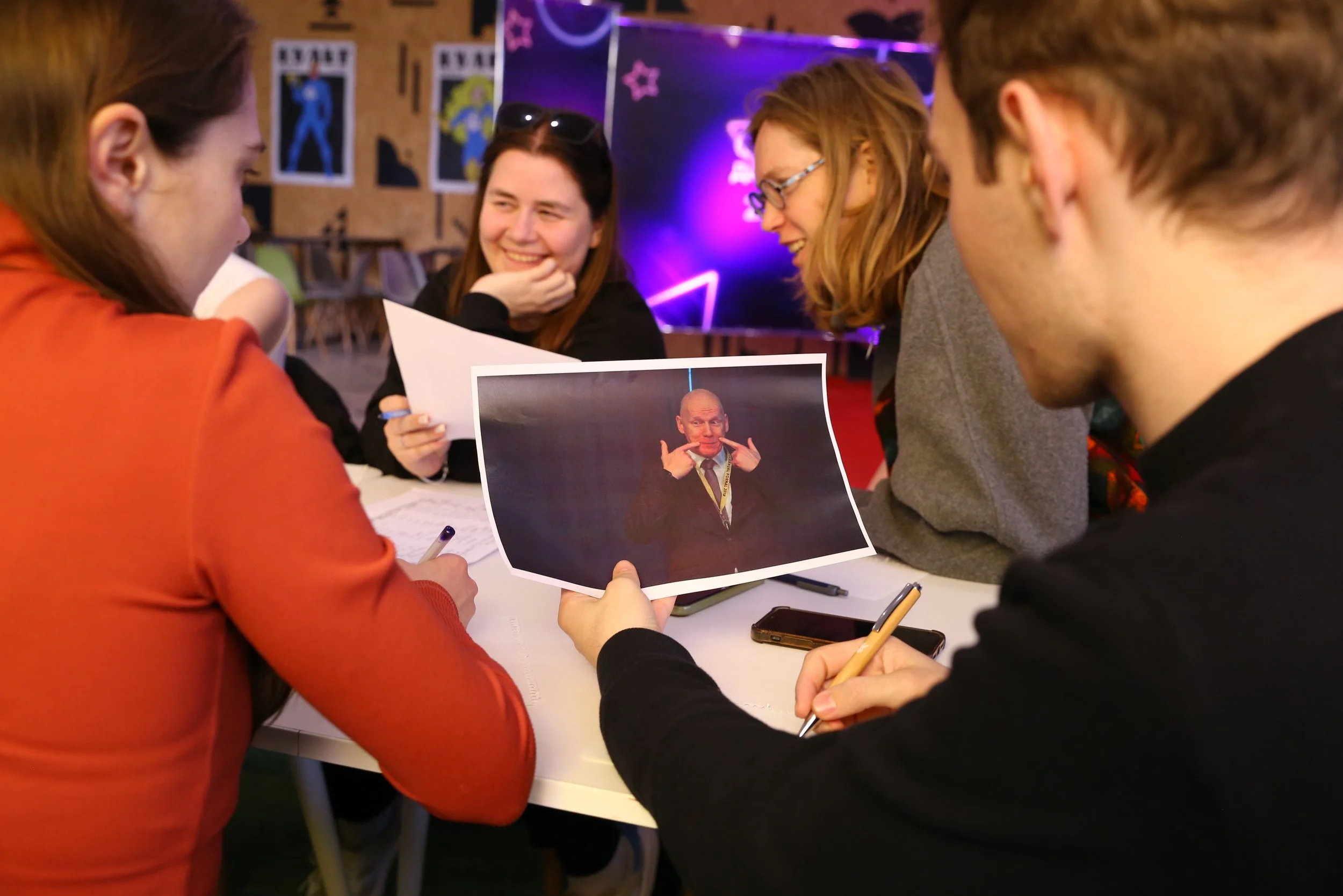 Group of five people sitting around a table, looking at a photo of a man making a gesture with his hands. They are smiling and appear to be having a fun discussion.