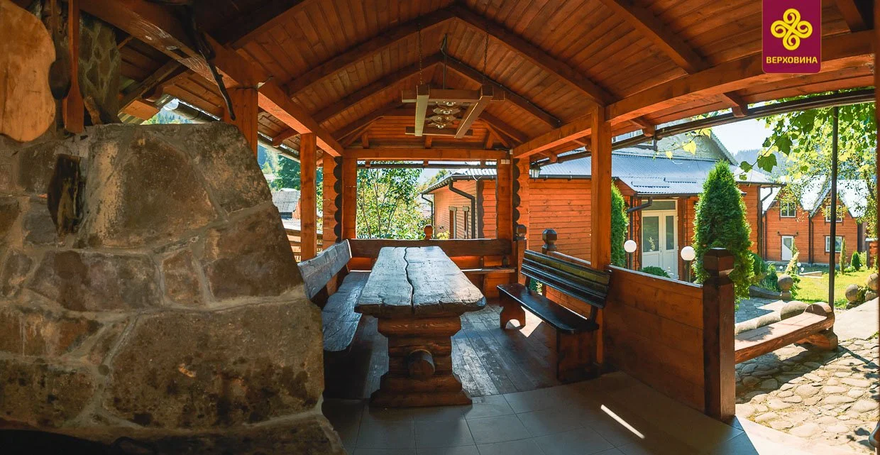 A covered outdoor porch area with a rustic wooden table and benches, stone fireplace on the left, and a garden with trees and a pathway outside.