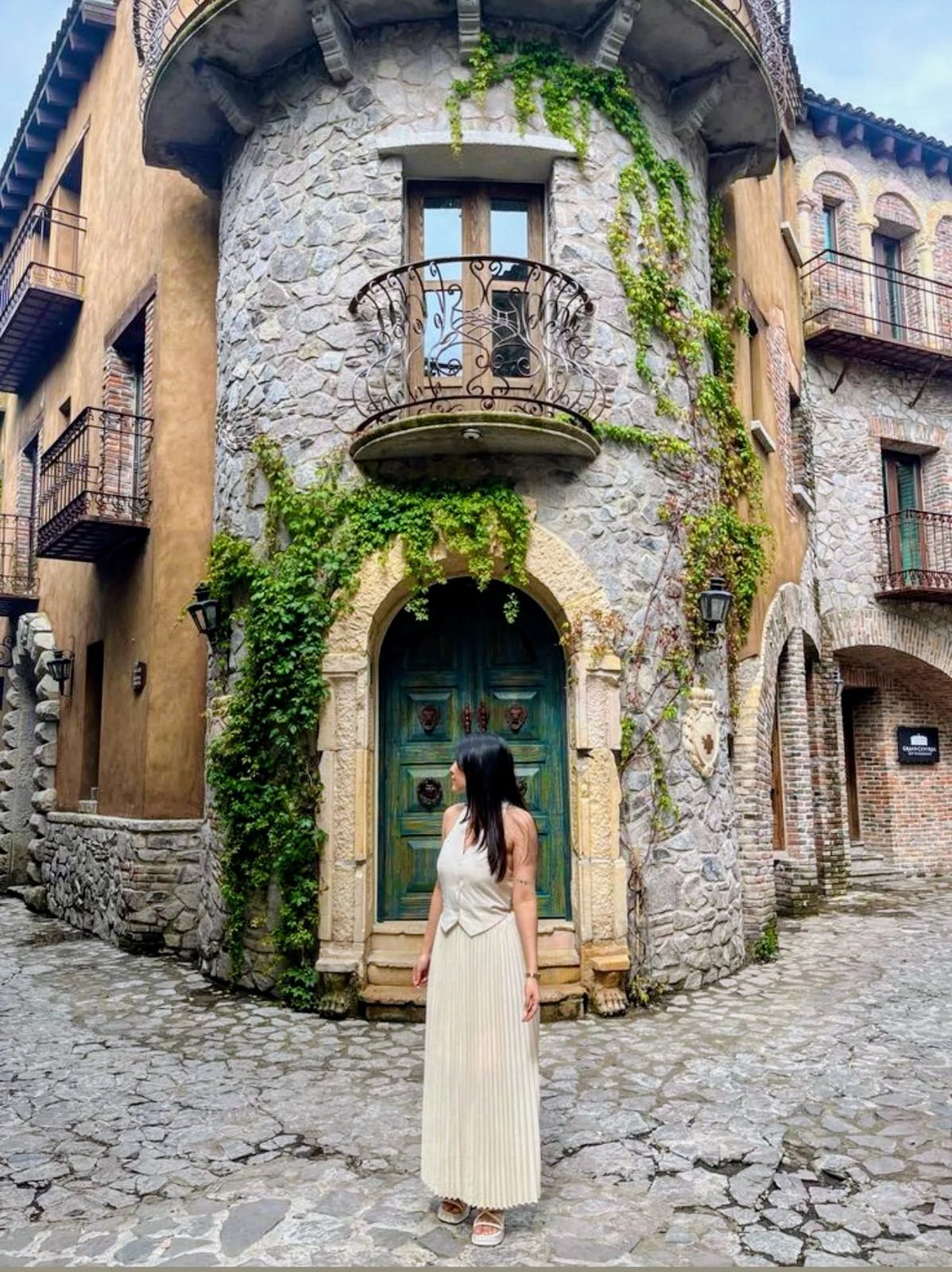 Una mujer de cabello oscuro y largo vestida con un largo vestido blanco de pie frente a una casa de estilo rústico con paredes de piedra y ventanas con balcón de hierro forjado en una calle de adoquines en Val'Quirico, Tlaxcala.