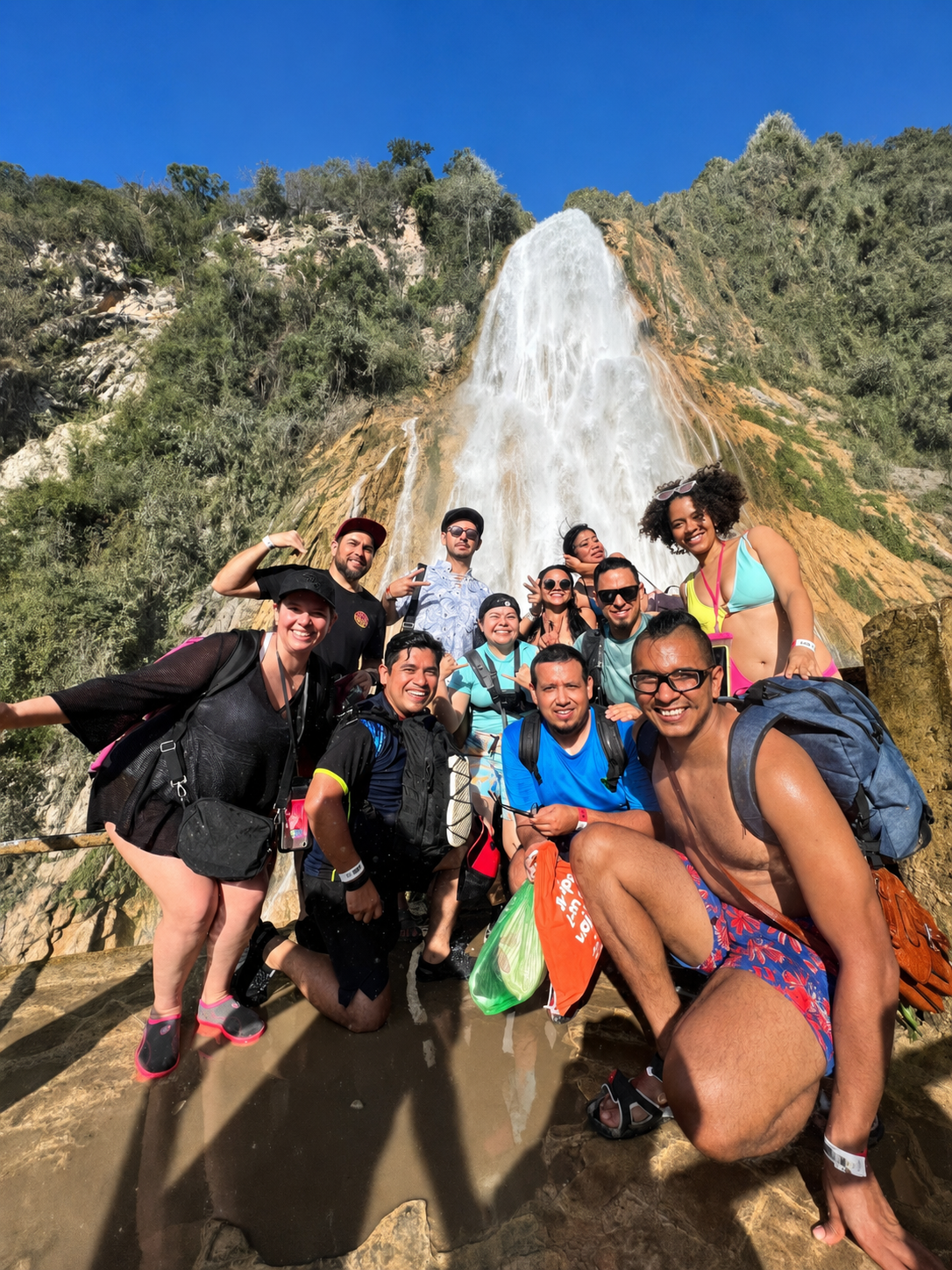 Grupo de personas sonriendo y posando en frente a la cascada del Chiflón en Chiapas, en un entorno natural, con árboles y cielo azul en el fondo.