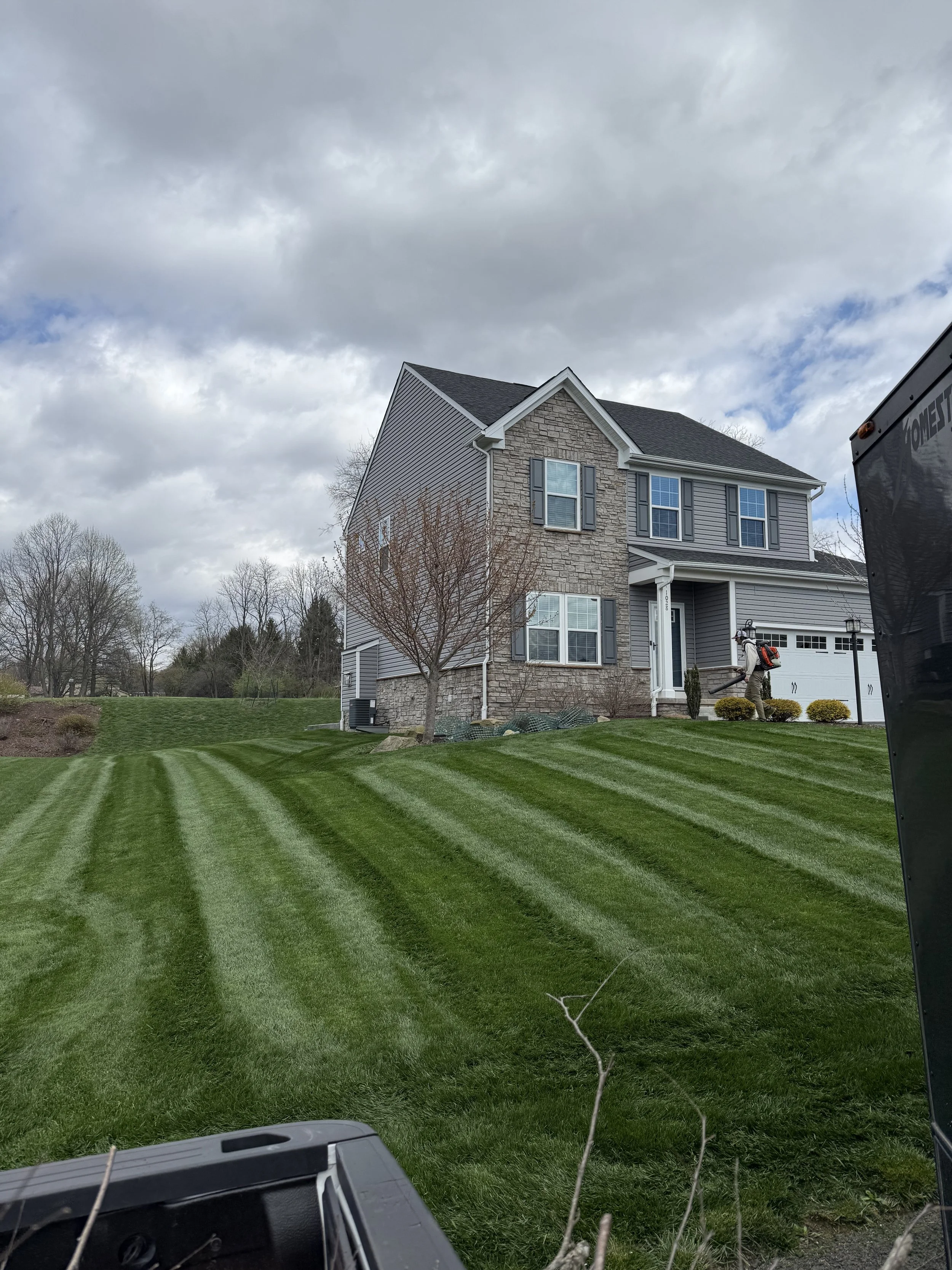 View of a well-maintained lawn in front of a two-story house with gray siding and stone accents, under a cloudy sky, with a person wearing a backpack and hoodie using a leaf blower near the porch.