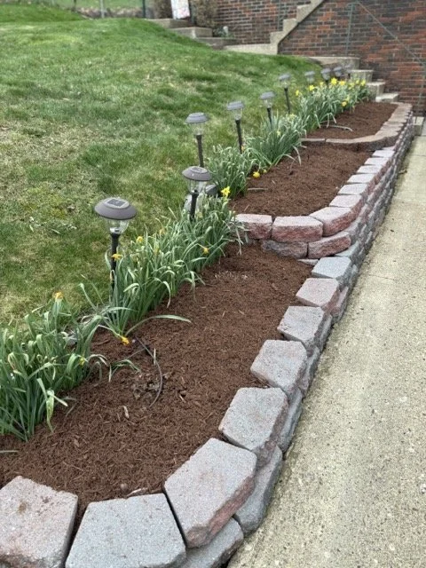 A landscaped garden bed with blooming yellow flowers and solar-powered garden lights, bordered by a mix of large and small bricks next to a sidewalk.