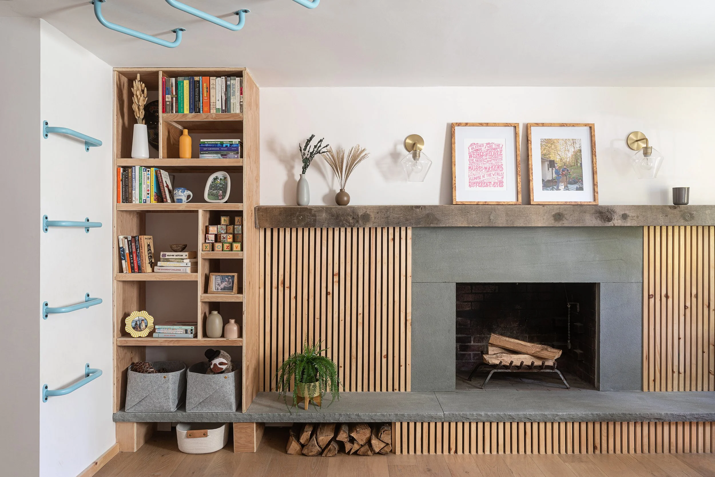 Living room with a fireplace, wooden shelving with books, photo frames, and decorative items, a potted fern, and mounted light fixtures.