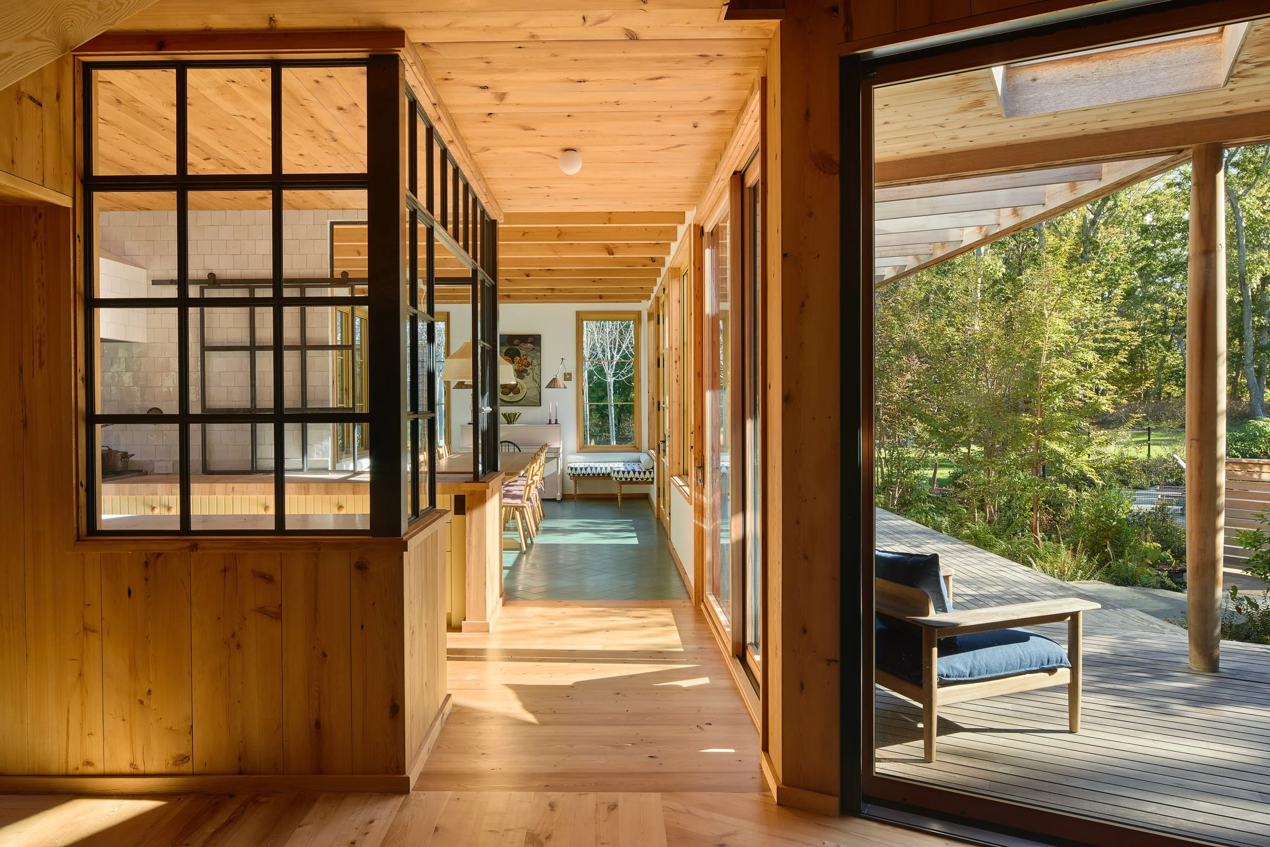 Interior view of a modern house with wooden walls, ceiling, and floor, leading to an outdoor deck and green foliage.