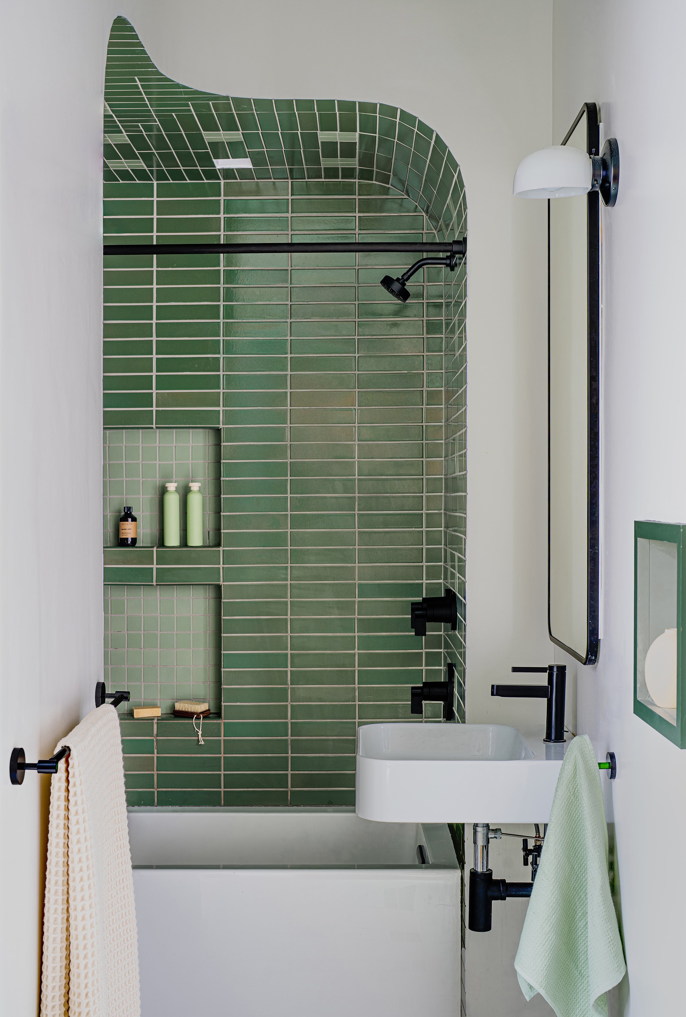 Bathroom with green tiled shower area, black fixtures, white sink, and white walls with black towel rack and a wall-mounted light fixture.