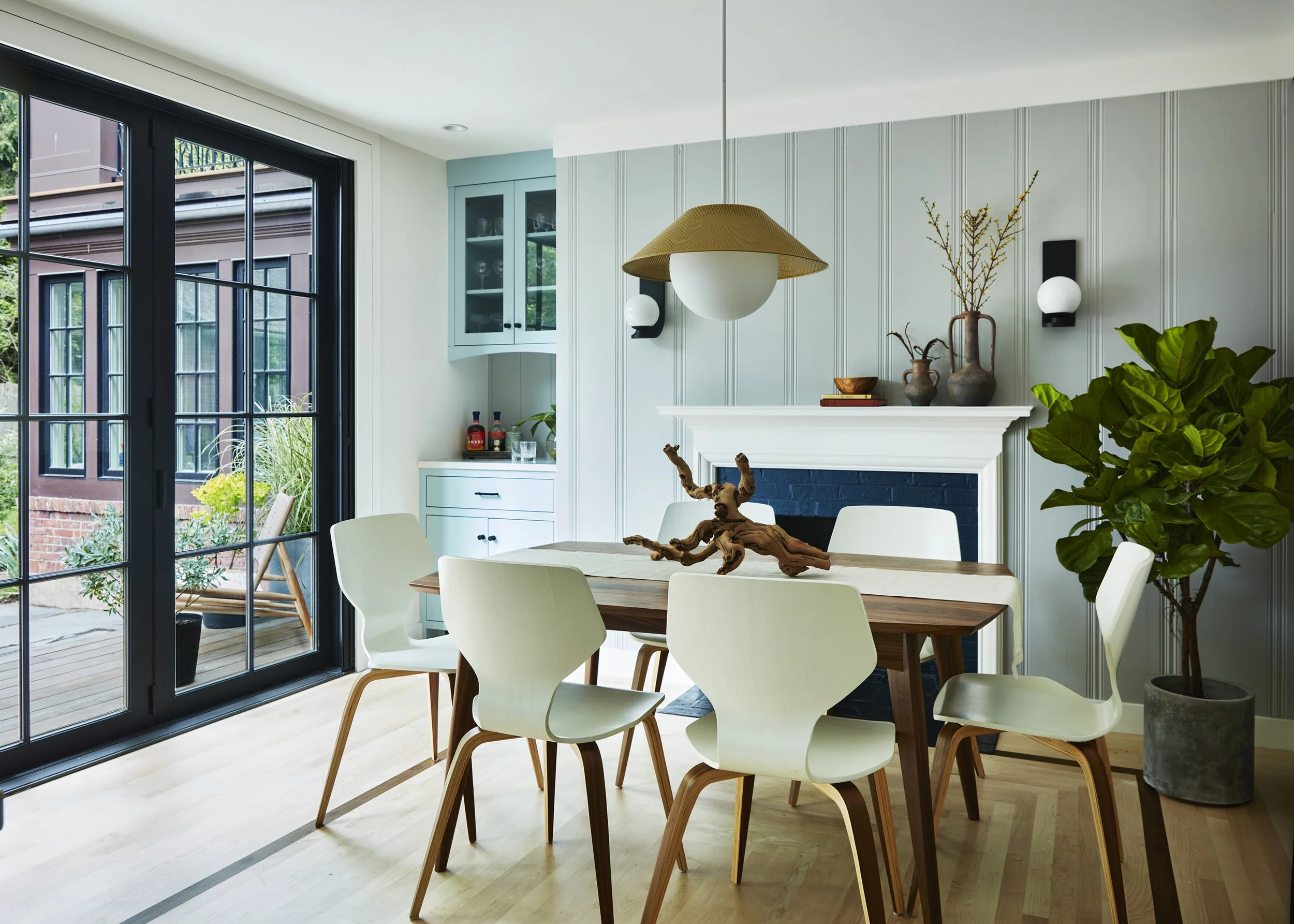 Modern dining room with light wood table, six white chairs, a fireplace with decorative vases and books, a potted plant, and large glass sliding doors leading outdoors.