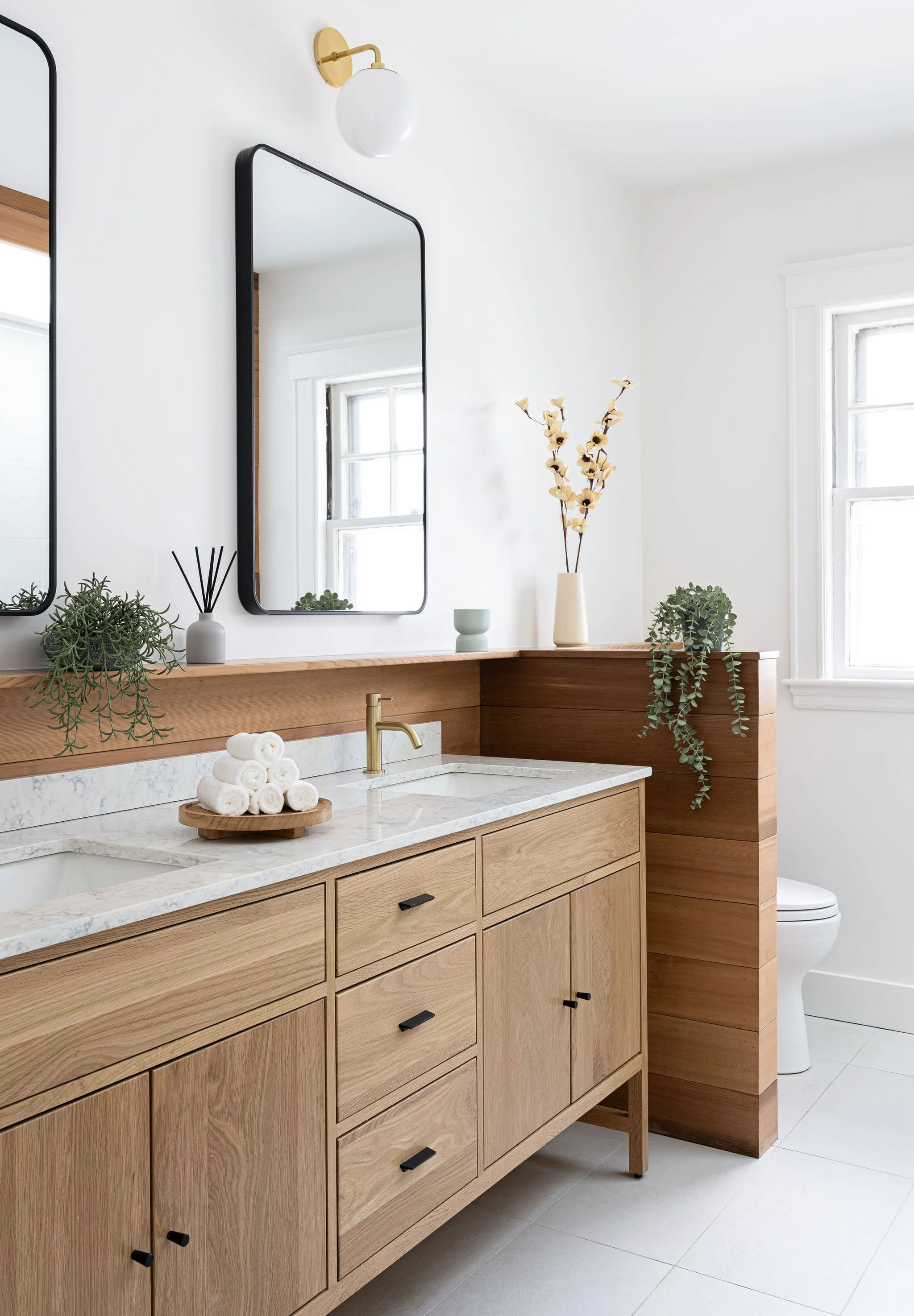 Modern bathroom with wooden vanity, white marble countertop, and two large mirrors. Decor includes potted plants, a vase with dried flowers, and a stack of rolled towels. Natural light from windows illuminates the space.