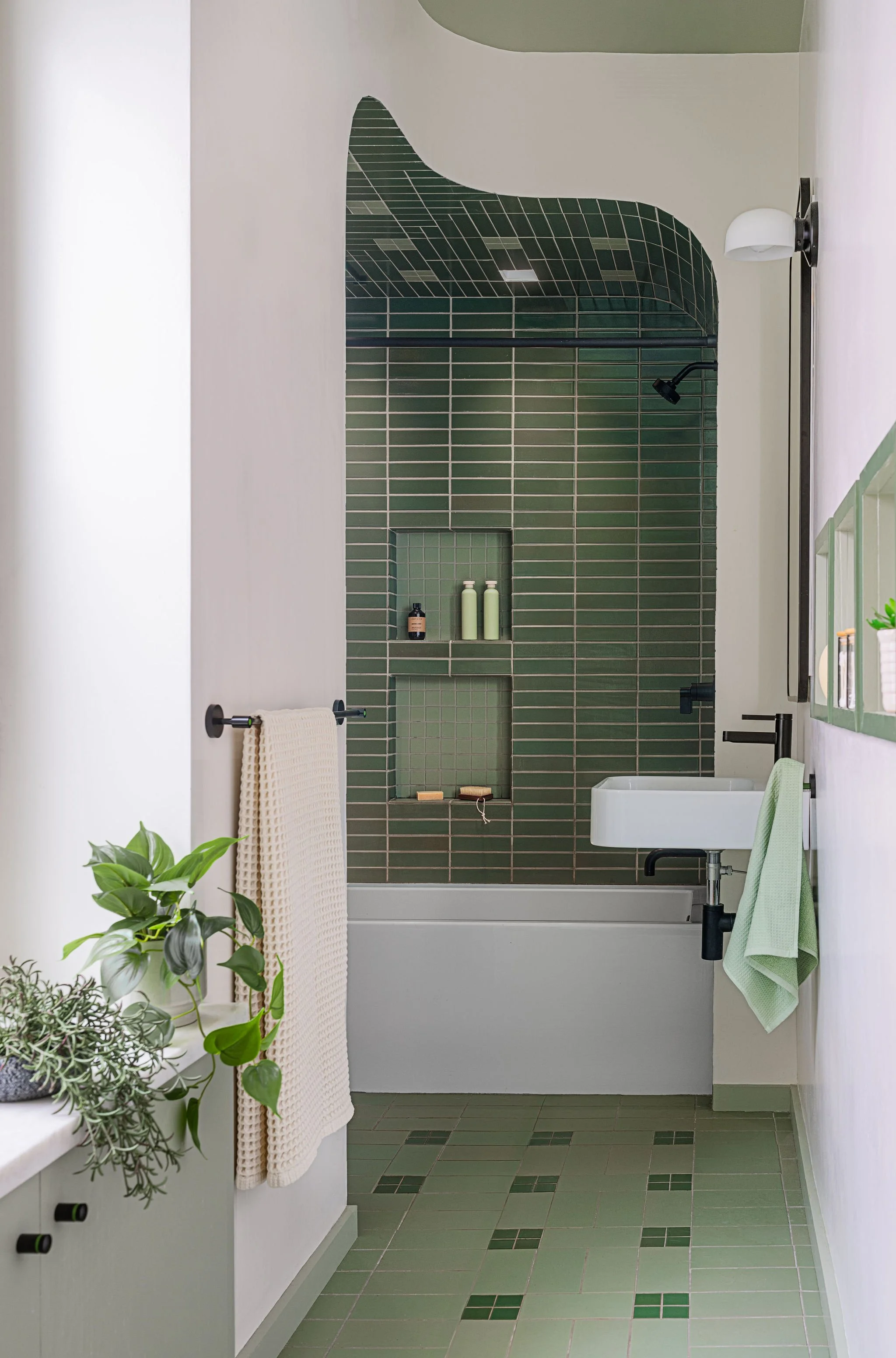 A modern bathroom with a green-tiled shower area, small shelf with toiletries, white bathtub, and a green and white color scheme, including plants and hand towels.