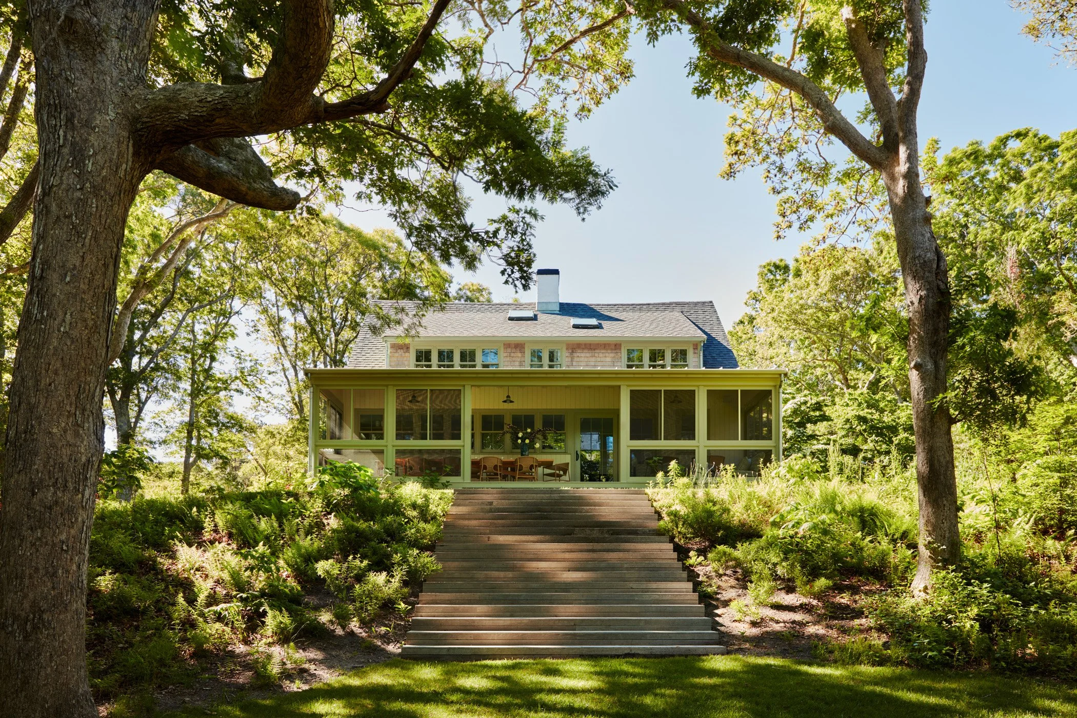 A house with a screened-in porch is accessed by a wide wooden staircase, set amidst lush greenery and tall trees with a clear blue sky overhead.