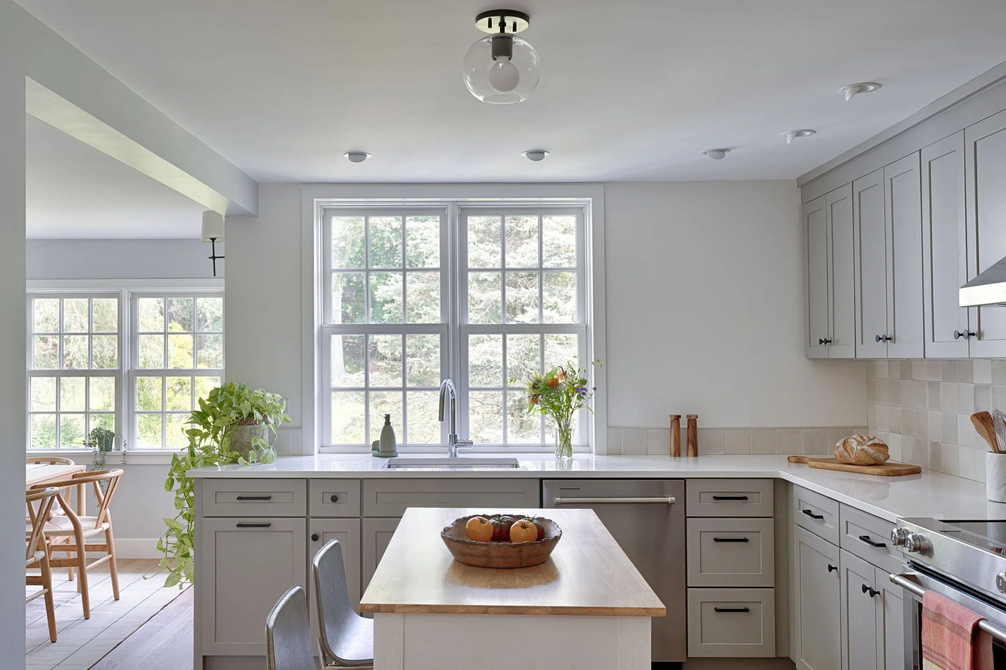 Bright kitchen with white cabinets, a central island, and large windows bringing in natural light. On the island, there is a fruit bowl with apples, and on the countertop, a loaf of bread. There are potted plants and a vase of flowers near the sink.
