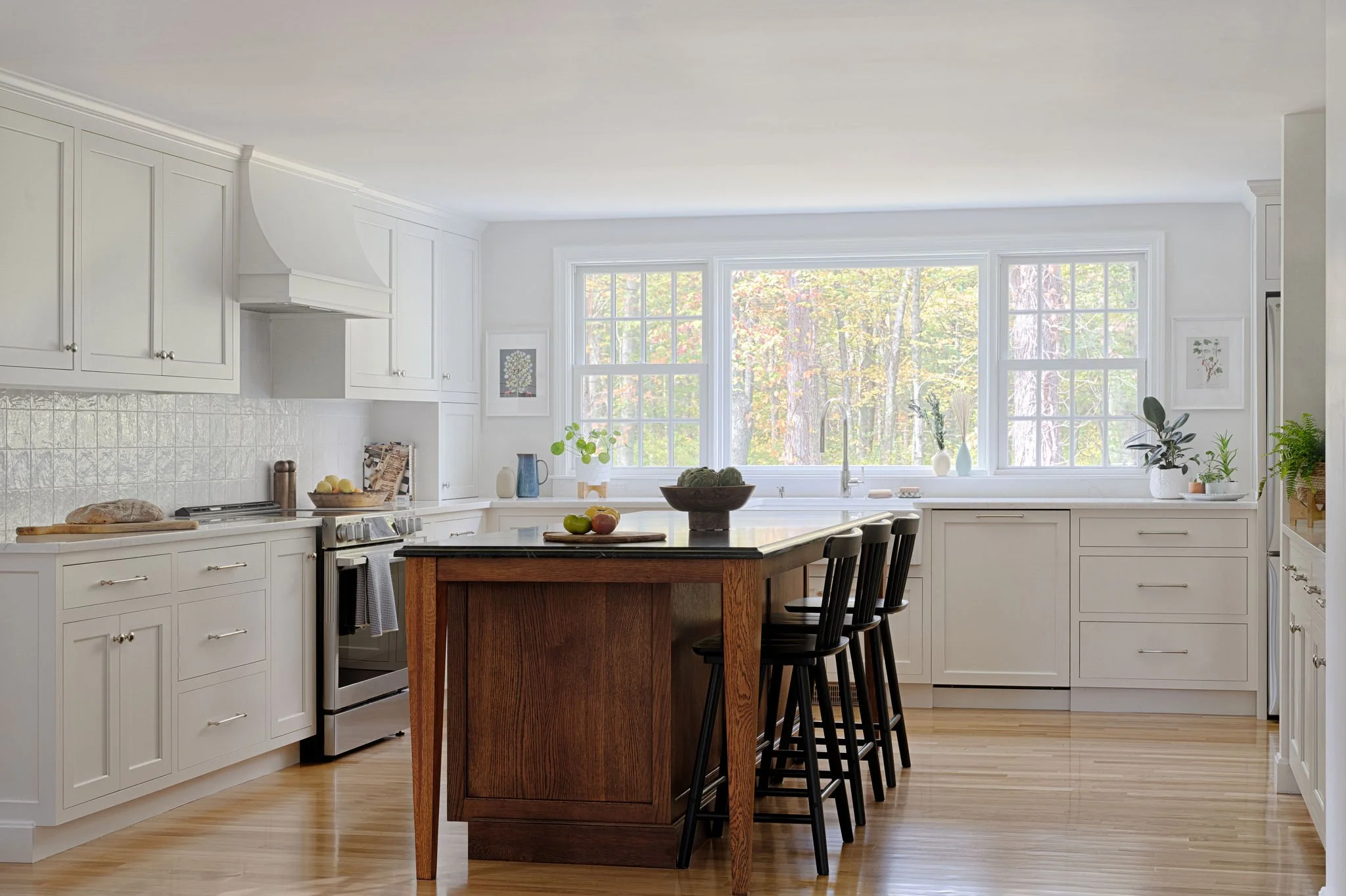 Bright kitchen with white cabinets, a large window, a black countertop island, black bar stools, and wooden flooring, decorated with plants and artwork.