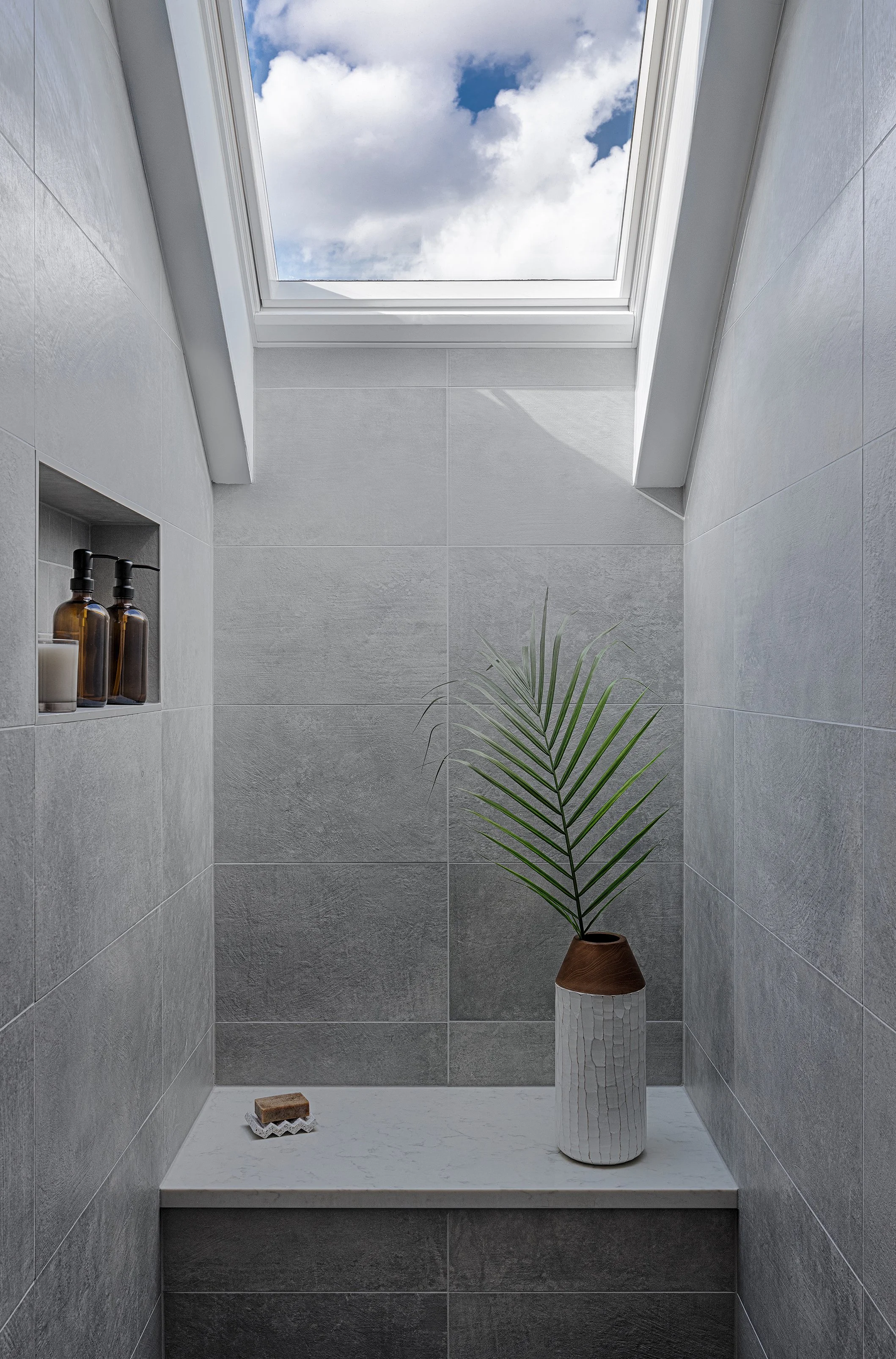 Shower stall with gray tiled walls, a skylight revealing a partly cloudy sky, a small niche with bottles and a candle, a potted palm plant, and a soap dish with a bar of soap.