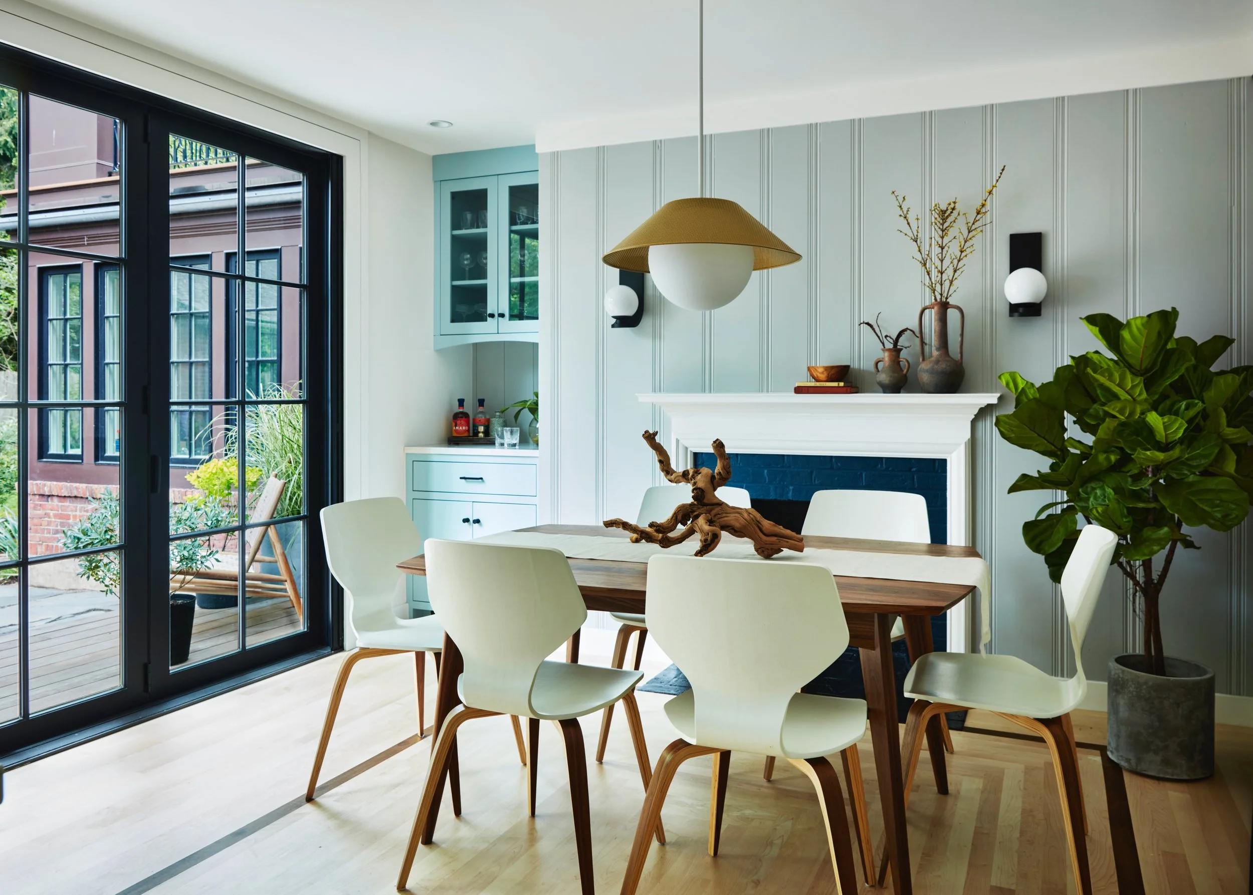Modern dining room with a wooden table, six white chairs, large window, potted plant, and decorative vases and dishes on the mantel above a fireplace.